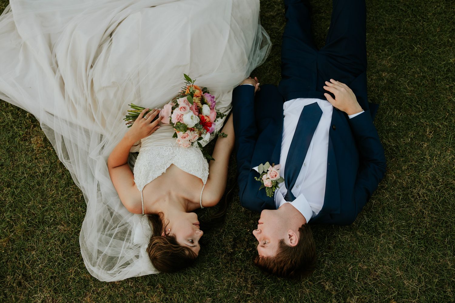 Bride and groom lying on green grass, bride holding colorful bouquet, wearing white gown and veil, groom in navy suit.
