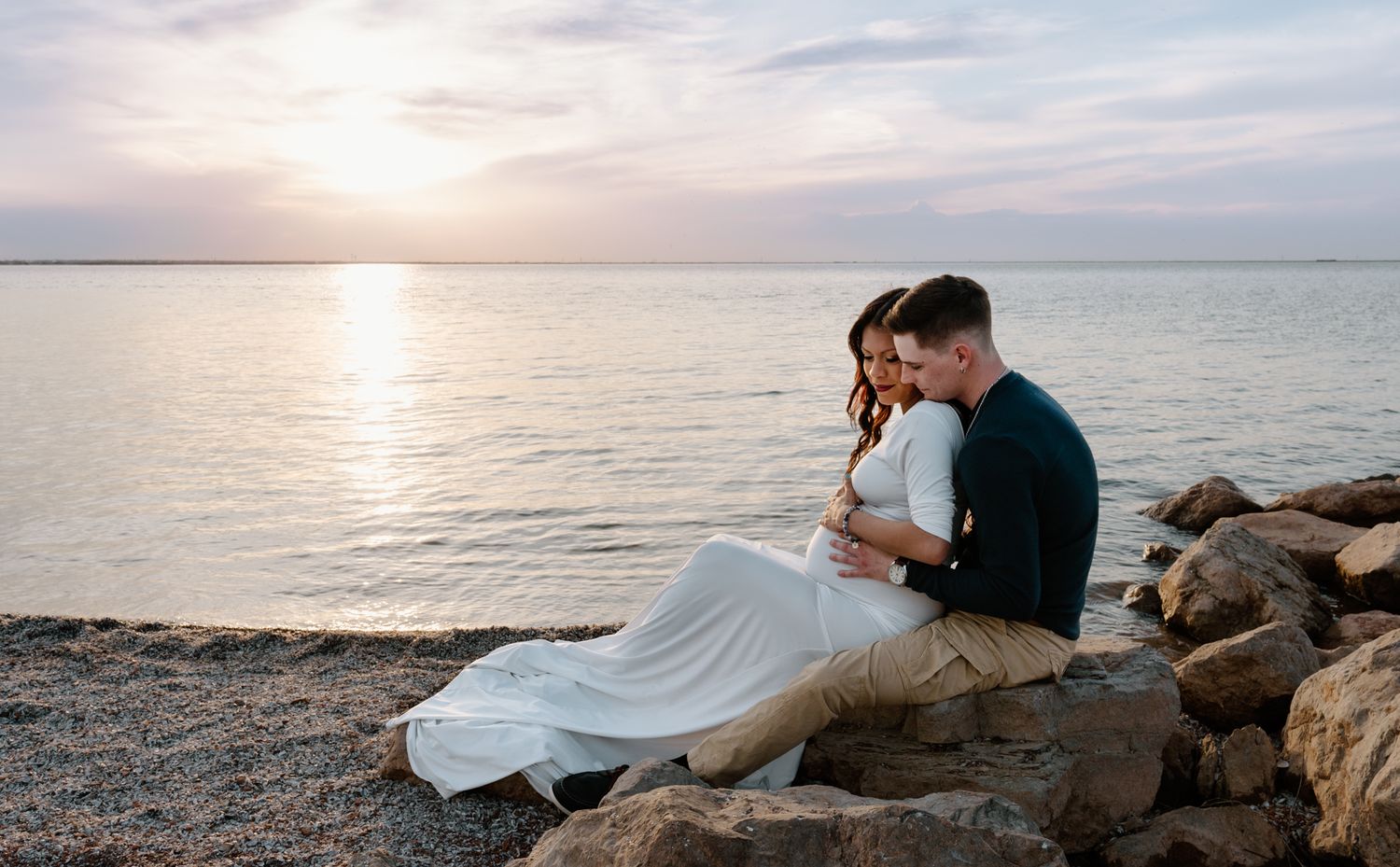 Couple sitting on rocks by the water at sunset, embracing tenderly during maternity photo session.