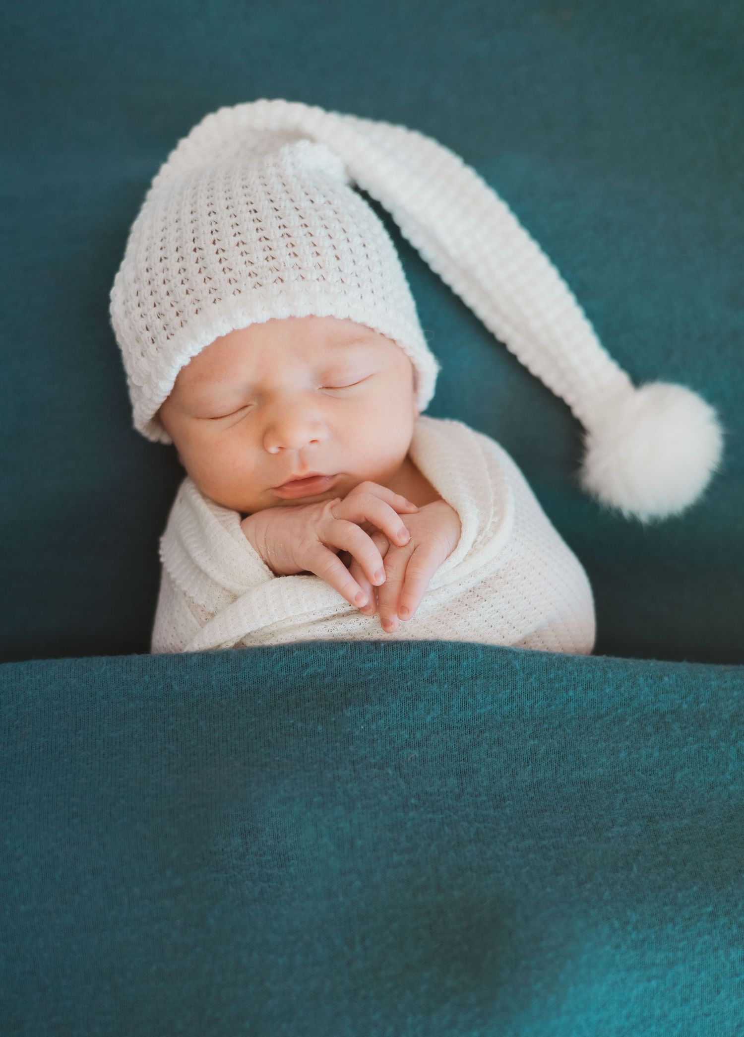 Newborn baby sleeping in white knit hat on teal blanket.