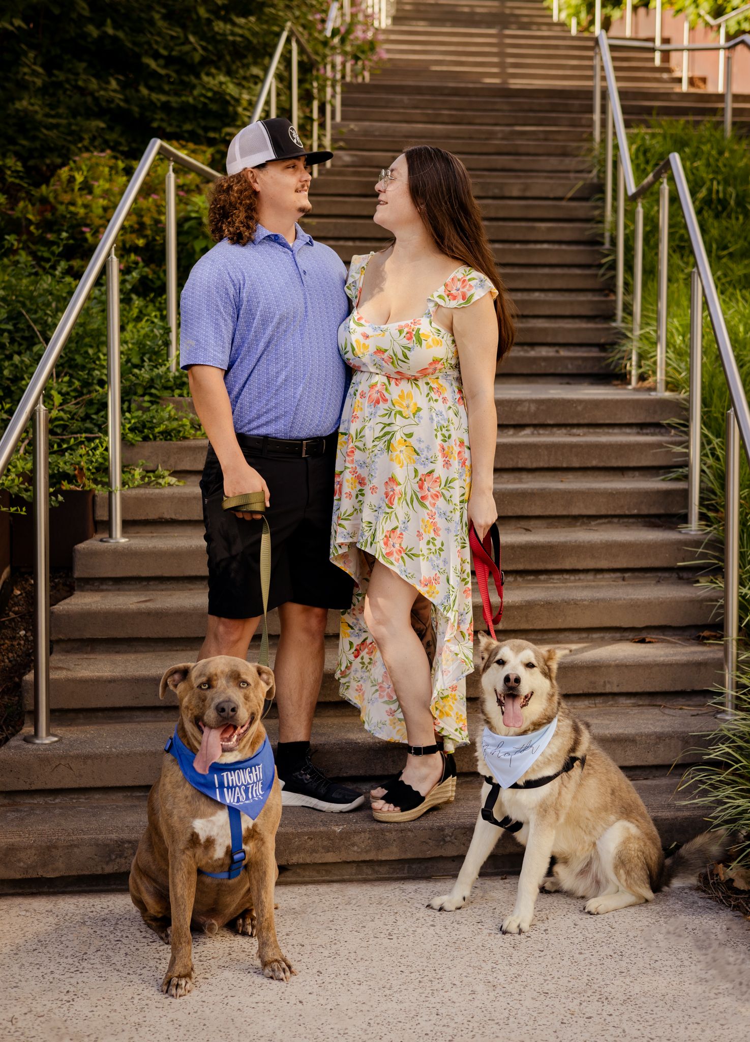 Young couple with two dogs wearing bandanas posing on outdoor steps.