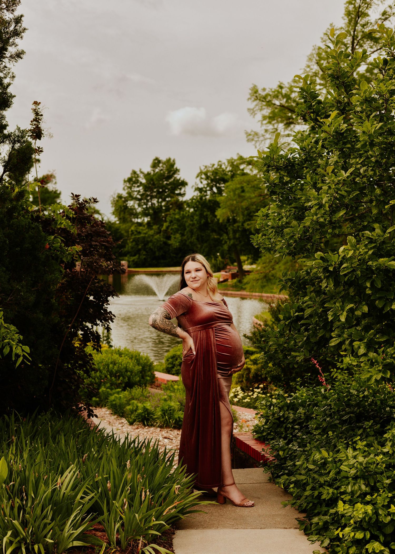 Pregnant woman in a burgundy maternity gown standing on a garden path with a fountain backdrop.