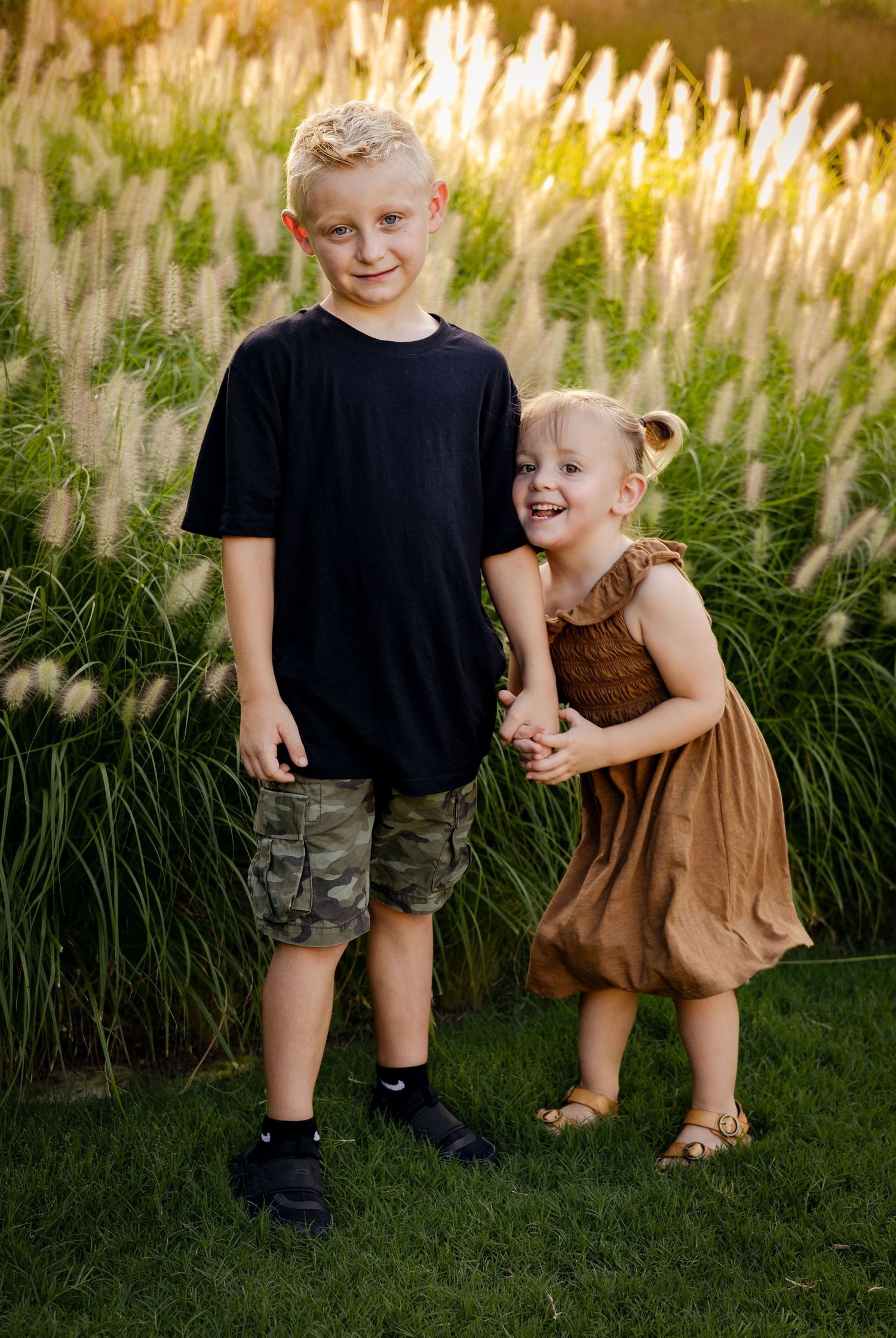 Two blonde siblings holding hands smiling in front of tall ornamental grass in warm golden hour light.