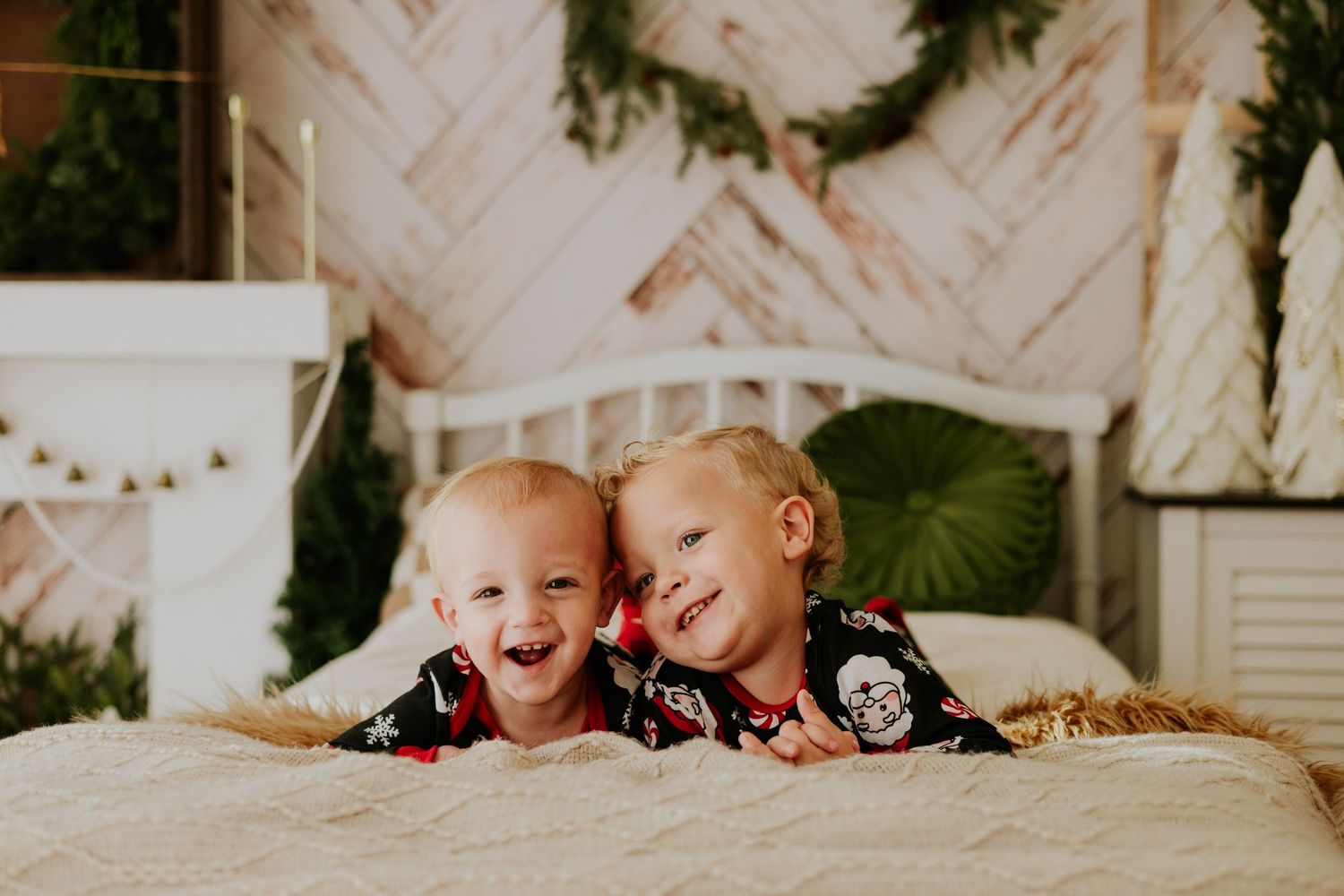 Two young blonde siblings in matching Christmas pajamas lying on a bed smiling and laughing together.