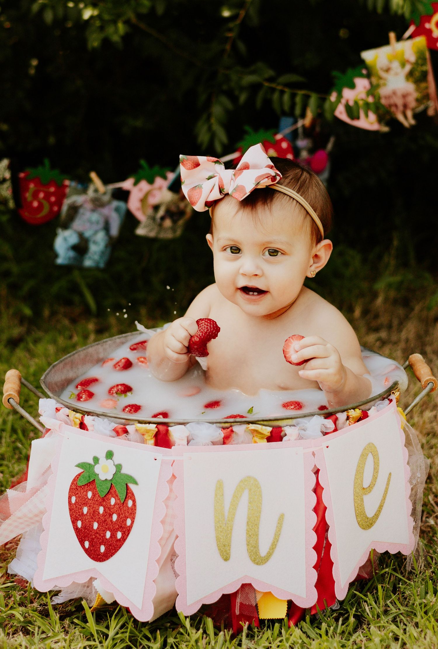 A smiling baby girl sits in a strawberry-themed milk bath tub decorated with a first birthday banner.