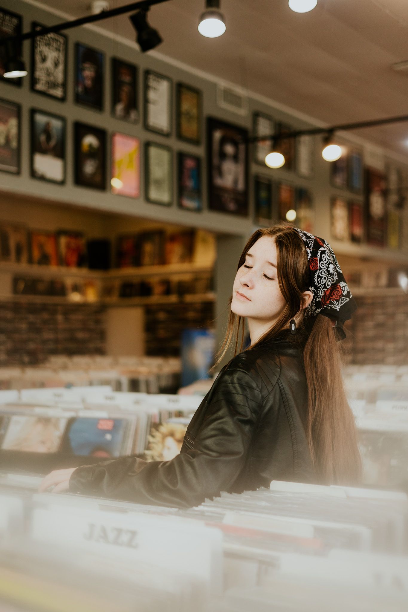 Young woman in leather jacket browsing jazz vinyl records in a cozy music store with album art on walls.