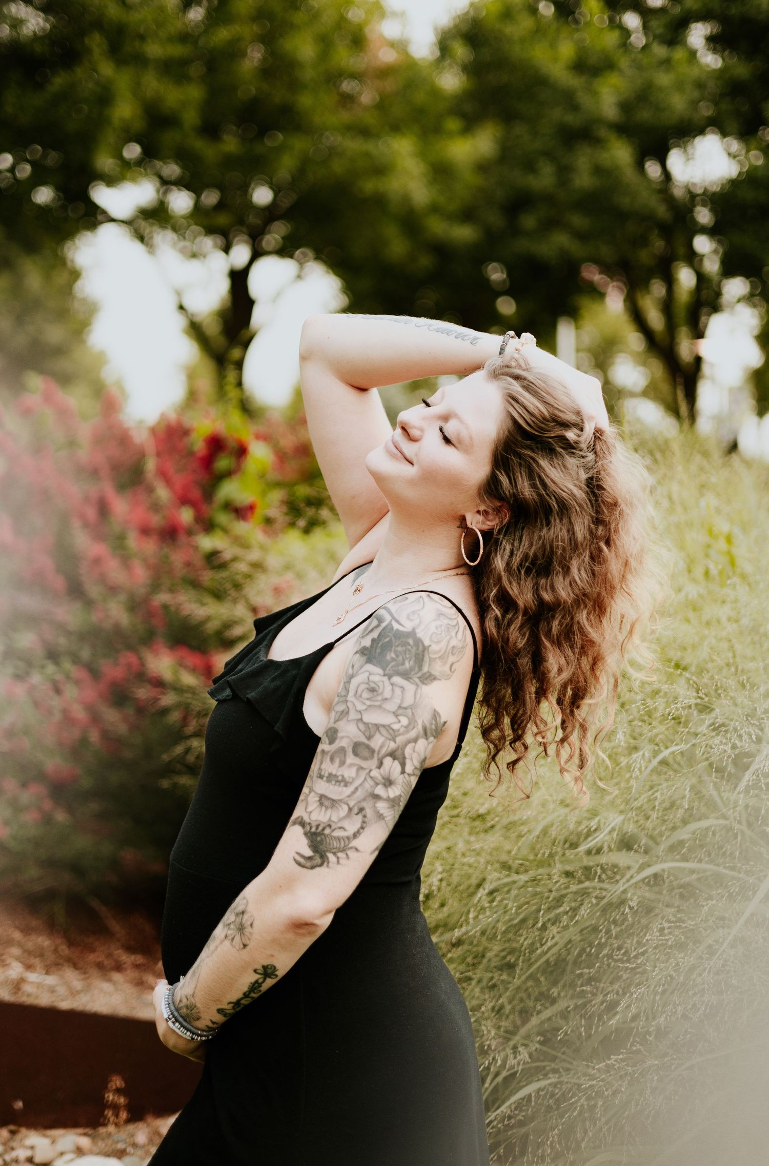A tattooed woman in a black dress poses with her hand in her hair among colorful garden flowers.