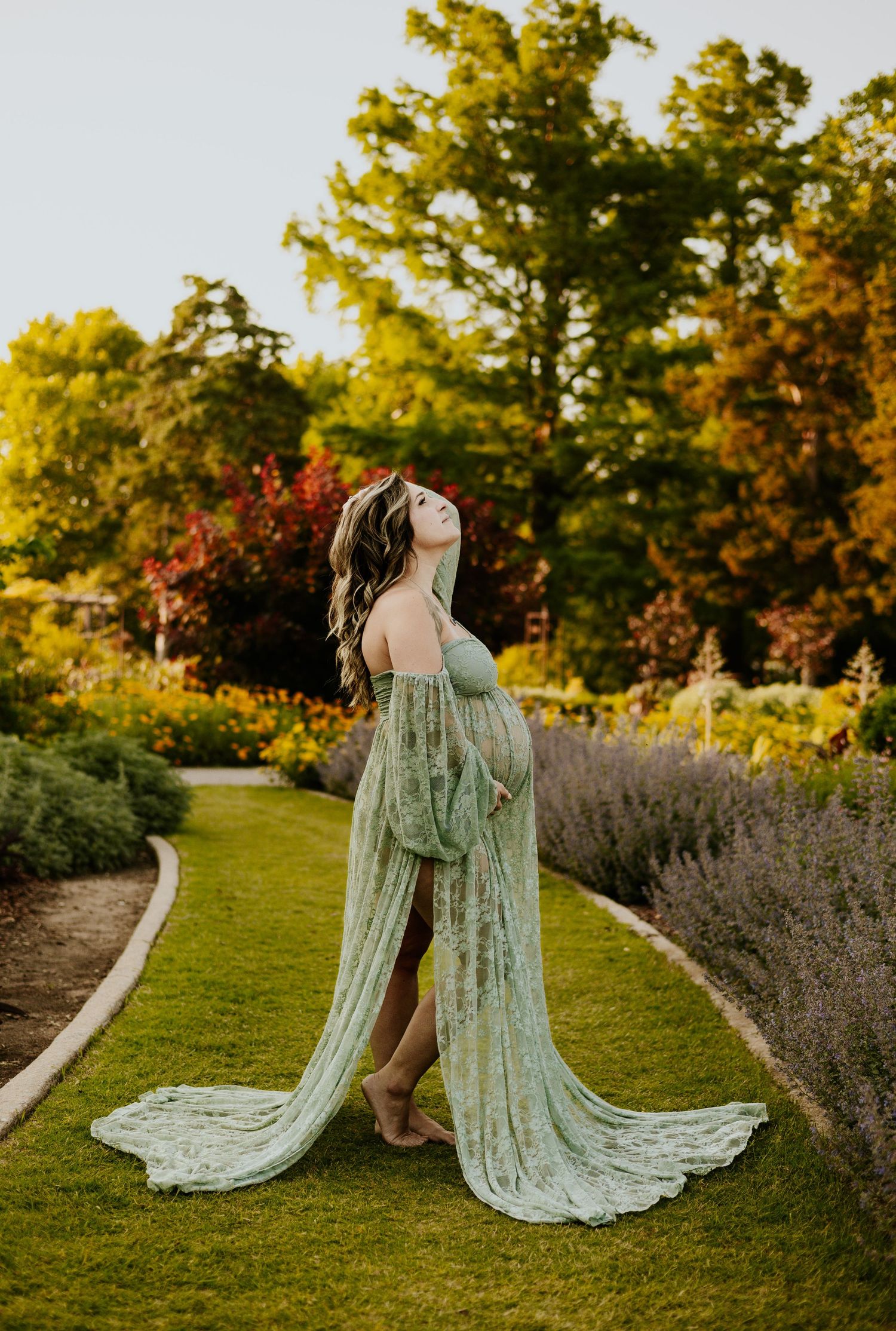 Pregnant woman in elegant sage green lace maternity gown posing in lush garden pathway at golden sunset.