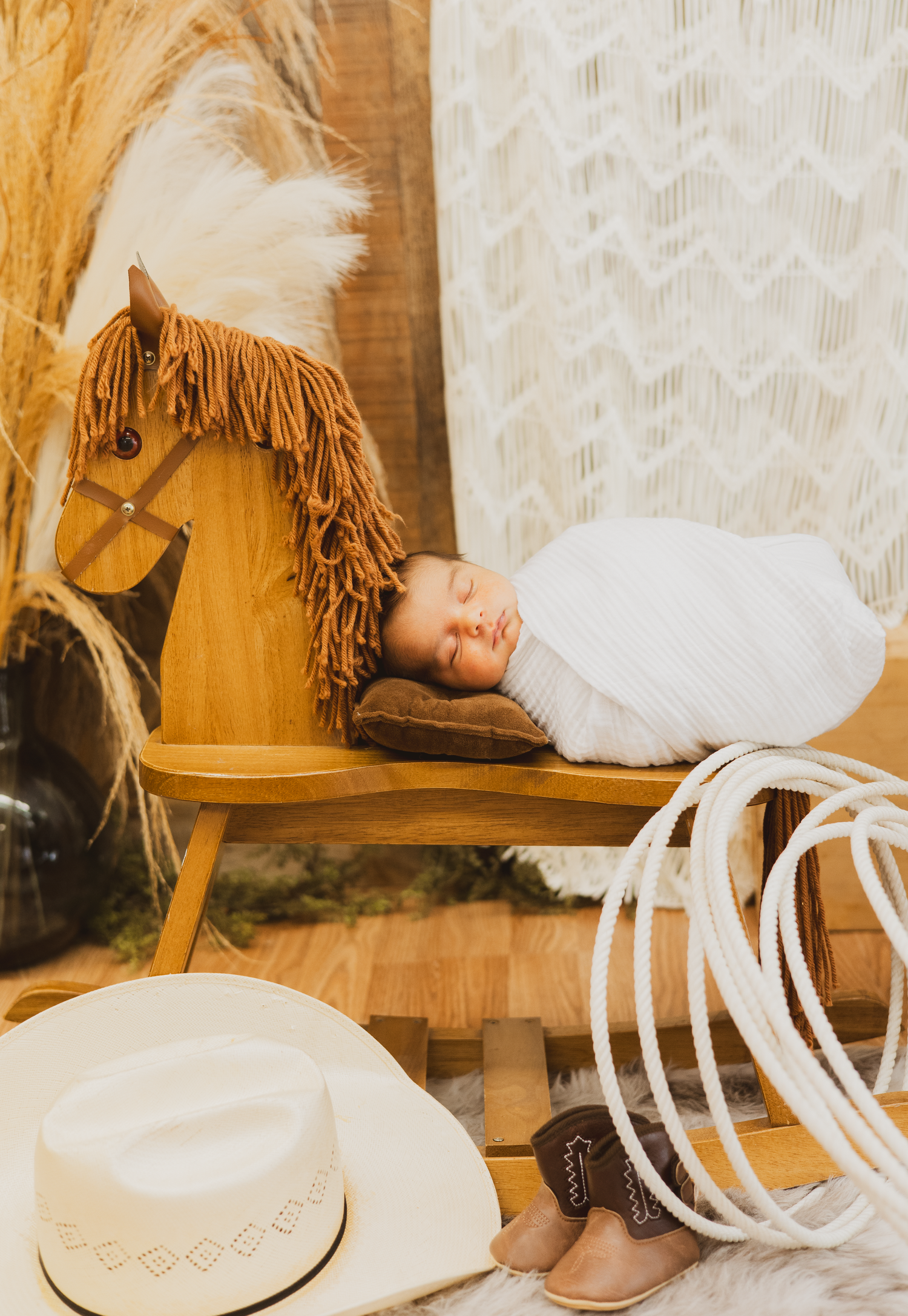 Newborn baby in white swaddle sleeping on wooden rocking horse with cowboy hat and boots.
