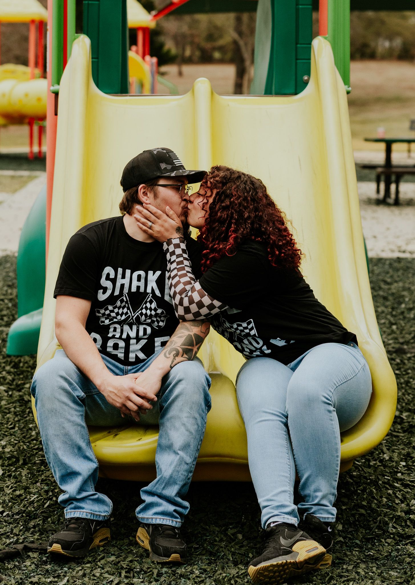 Couple kissing on a yellow playground slide, wearing matching Shake and Bake black t-shirts and jeans.