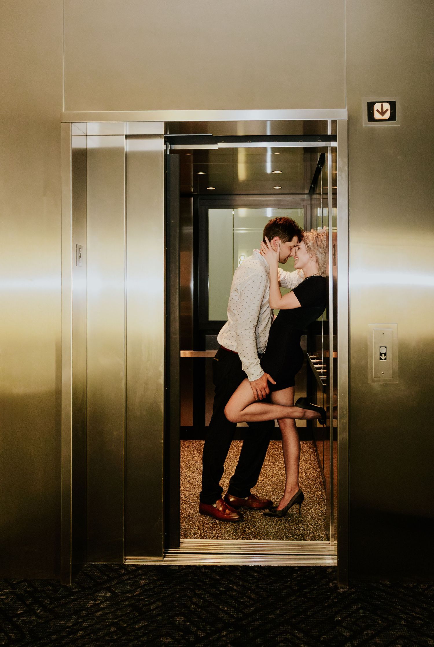 Couple sharing a passionate kiss inside a sleek silver elevator with open doors.