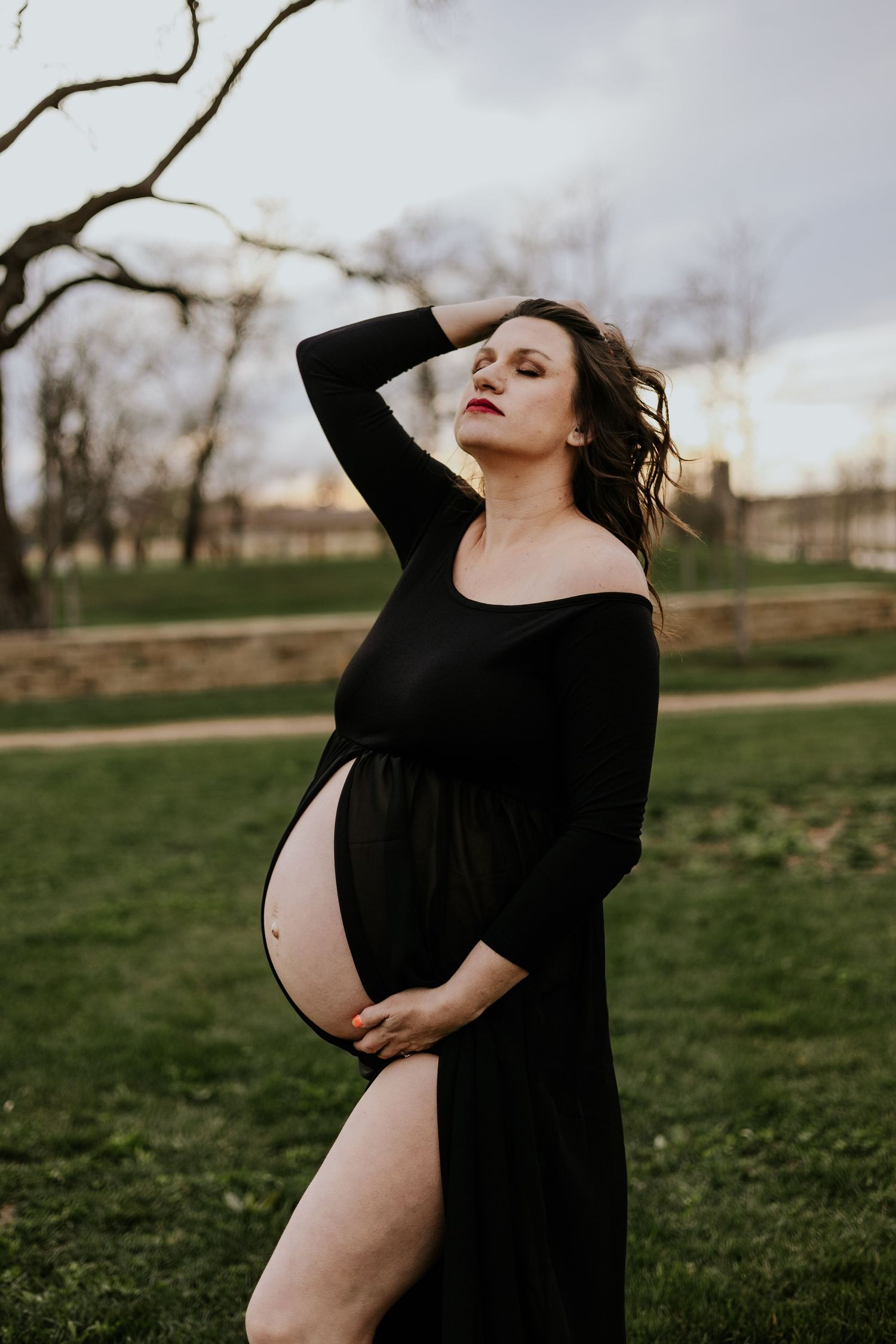 Pregnant woman in elegant black off-shoulder maternity dress poses gracefully in park at dusk.