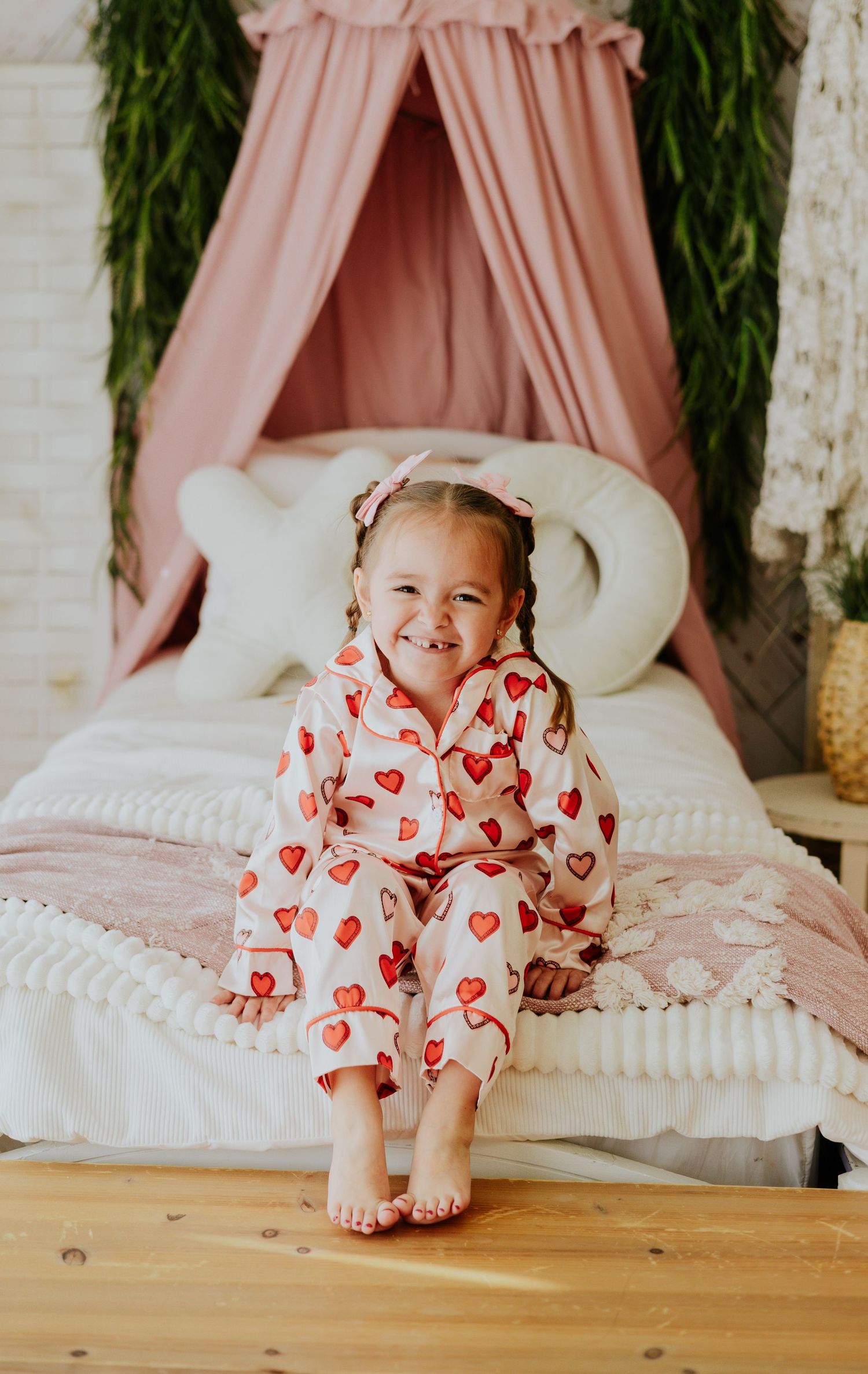 Adorable toddler girl in heart-print pajamas smiles while sitting on bed with pink canopy backdrop.