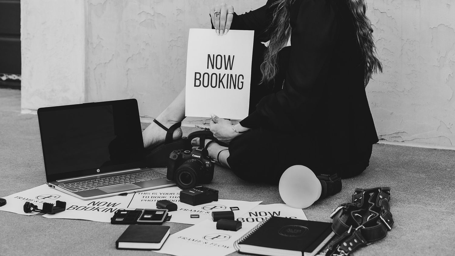 Woman sitting on floor holding a Now Booking sign surrounded by cameras, laptop, and photography equipment.