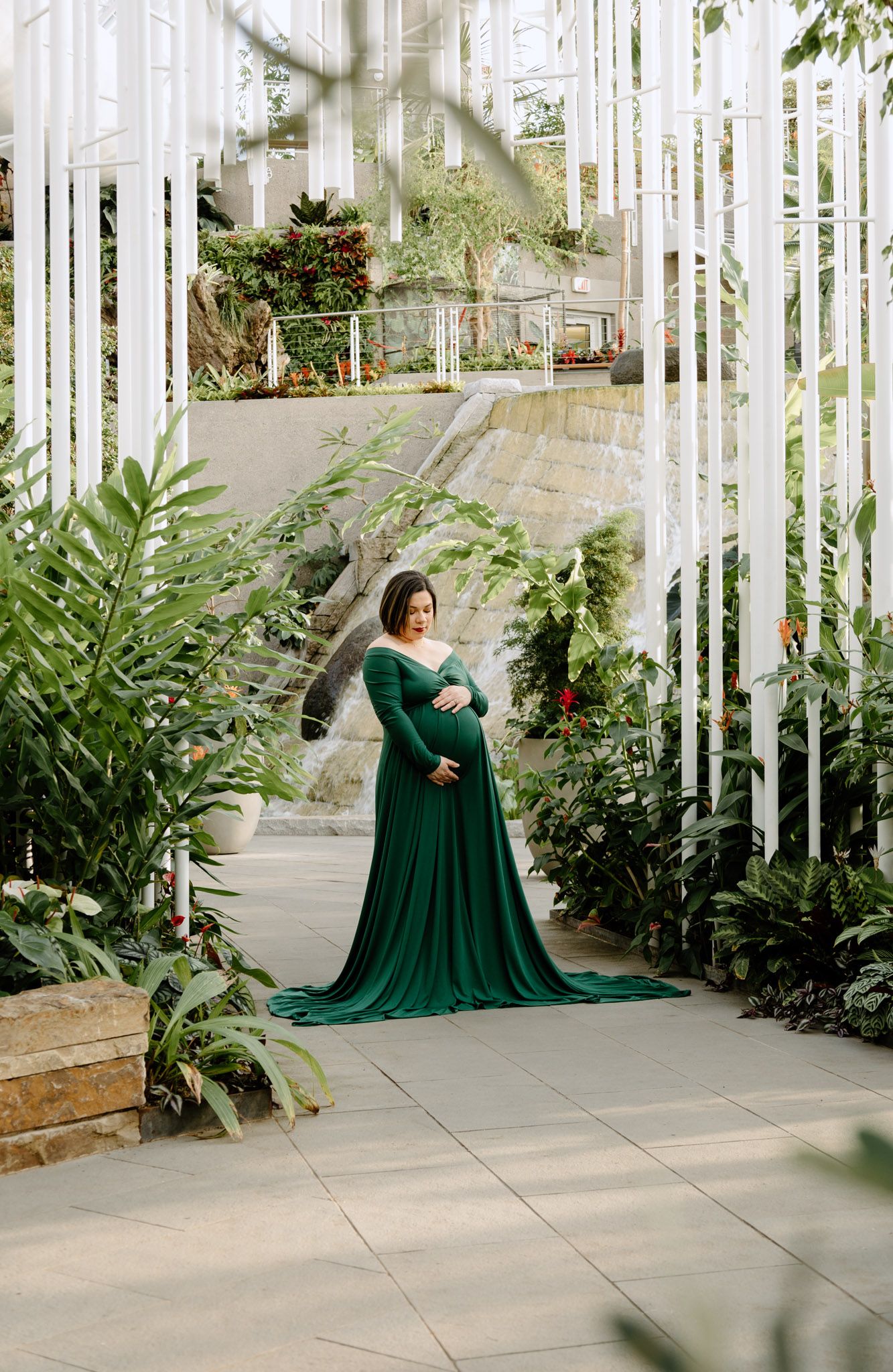 Pregnant woman in elegant emerald green gown standing among lush tropical plants in a greenhouse.