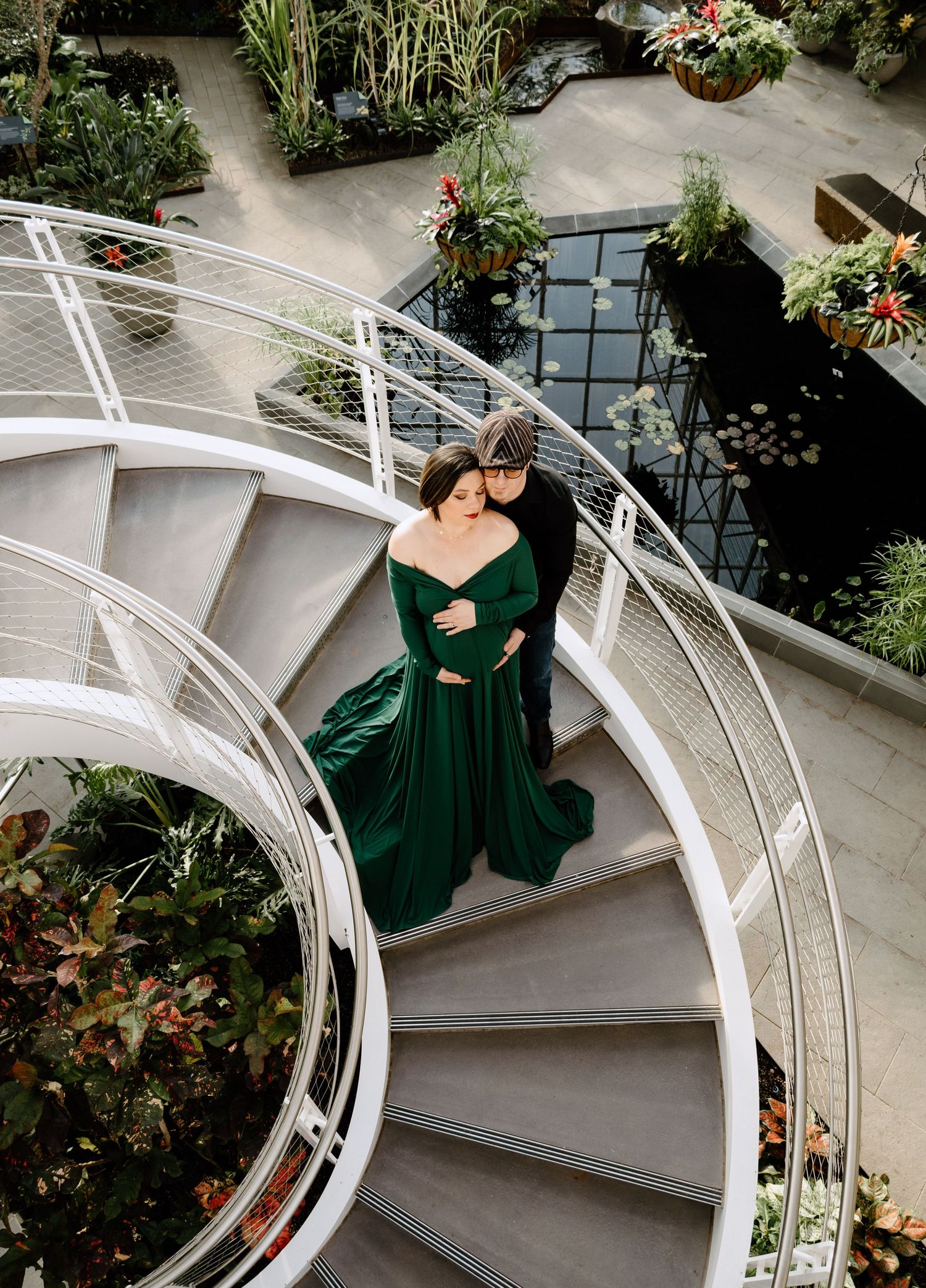 Aerial view of couple on spiral staircase, woman in emerald green maternity gown in a greenhouse.
