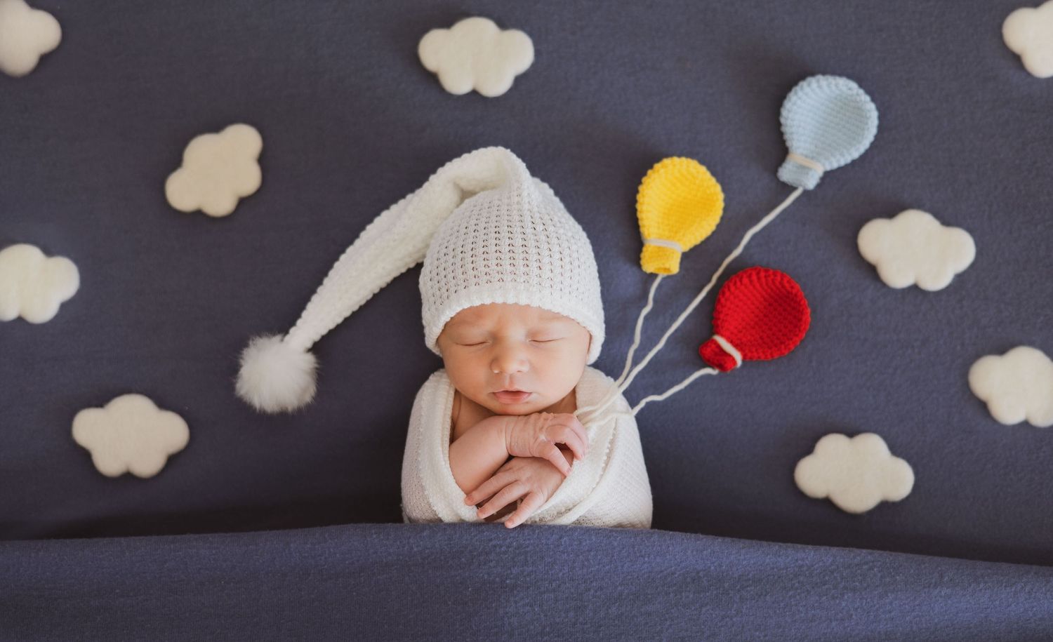 Newborn baby in white hat surrounded by cloud and balloon decor.