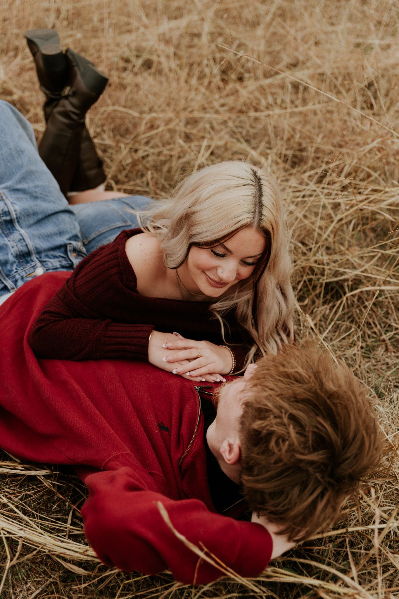 Couple lying in a golden field, woman in burgundy dress smiling softly at her partner in an intimate pose.