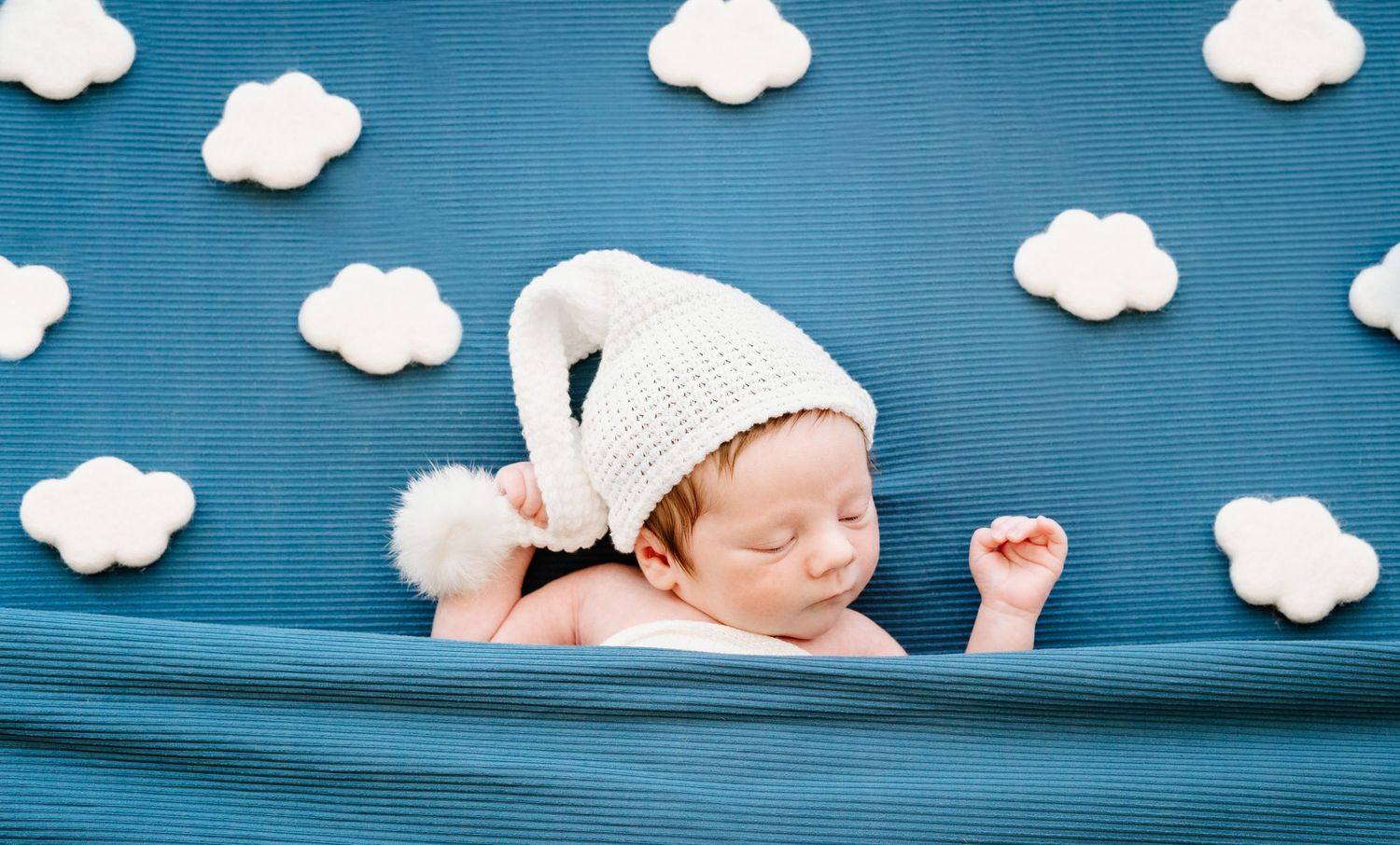 Sleeping newborn baby wearing a knit hat on a blue blanket with white cloud decorations.