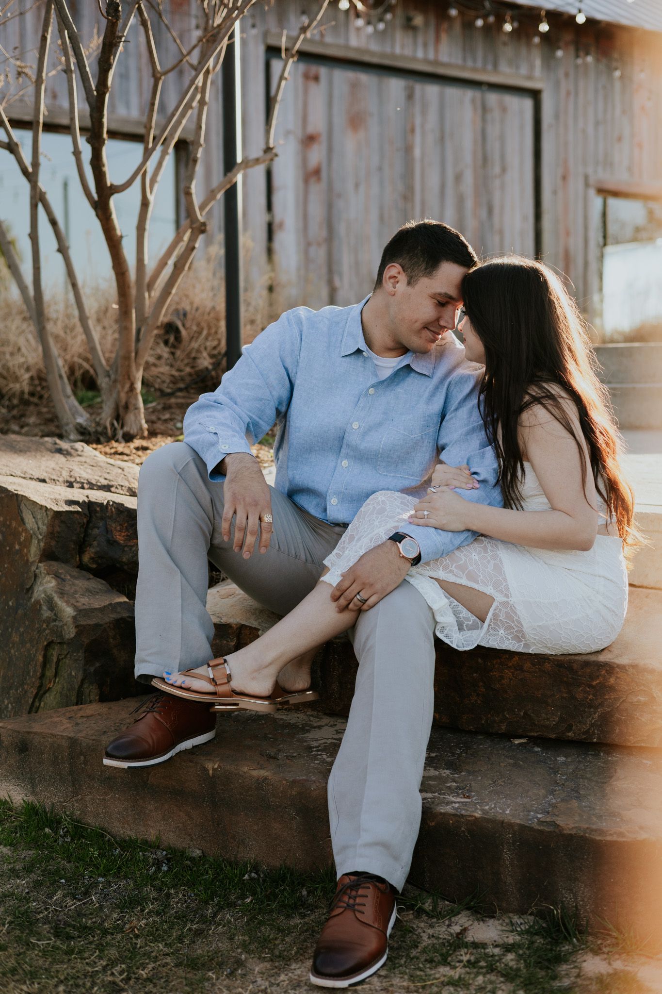 Couple sitting close on stone steps at golden hour, man in blue shirt, woman in white lace dress.