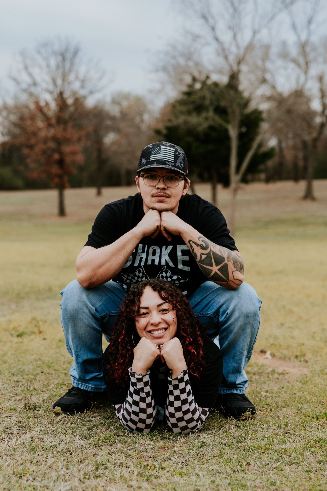 Man squatting behind woman lying on grass, both smiling for a playful couples photo in a park.
