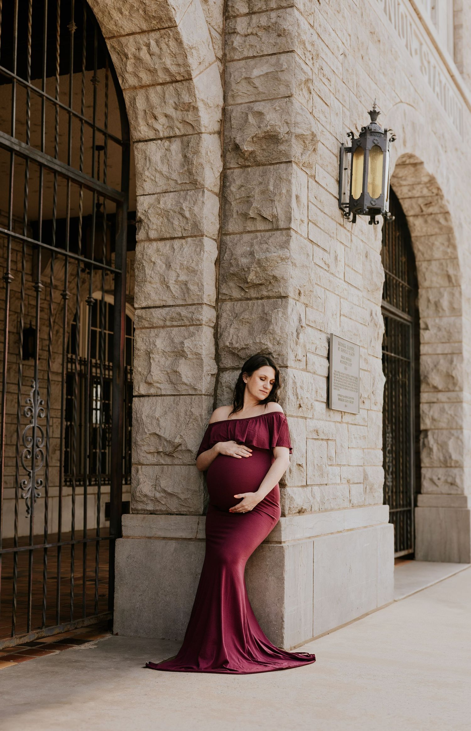 Pregnant woman in burgundy off-shoulder maternity gown posing by stone arch building.