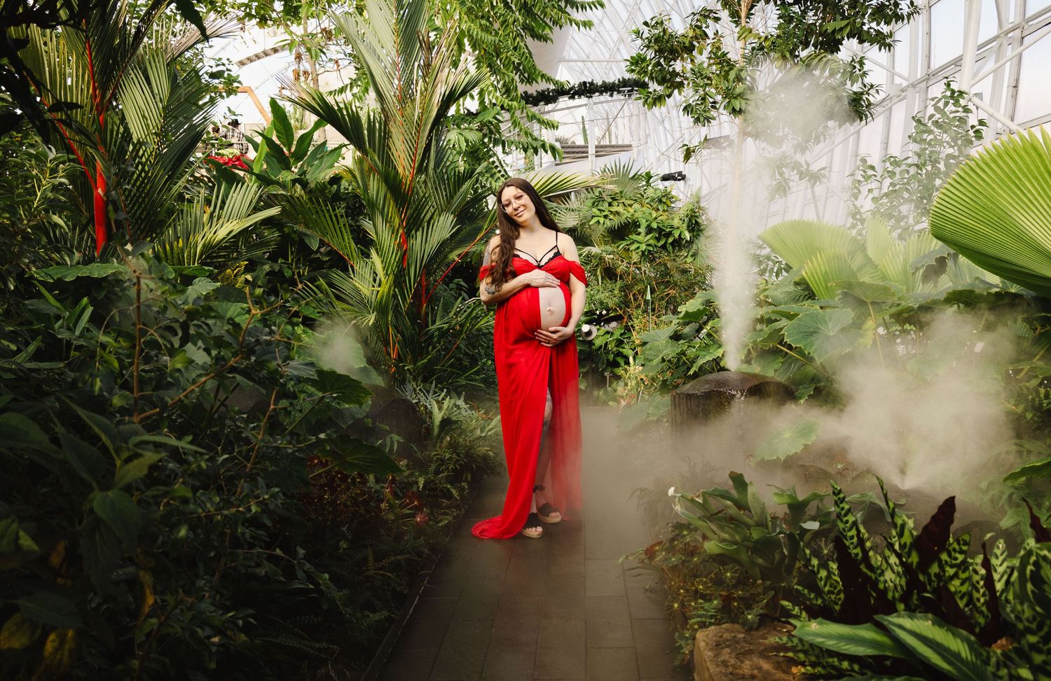 Expectant mother in a red outfit posing among lush tropical plants in a misty greenhouse.
