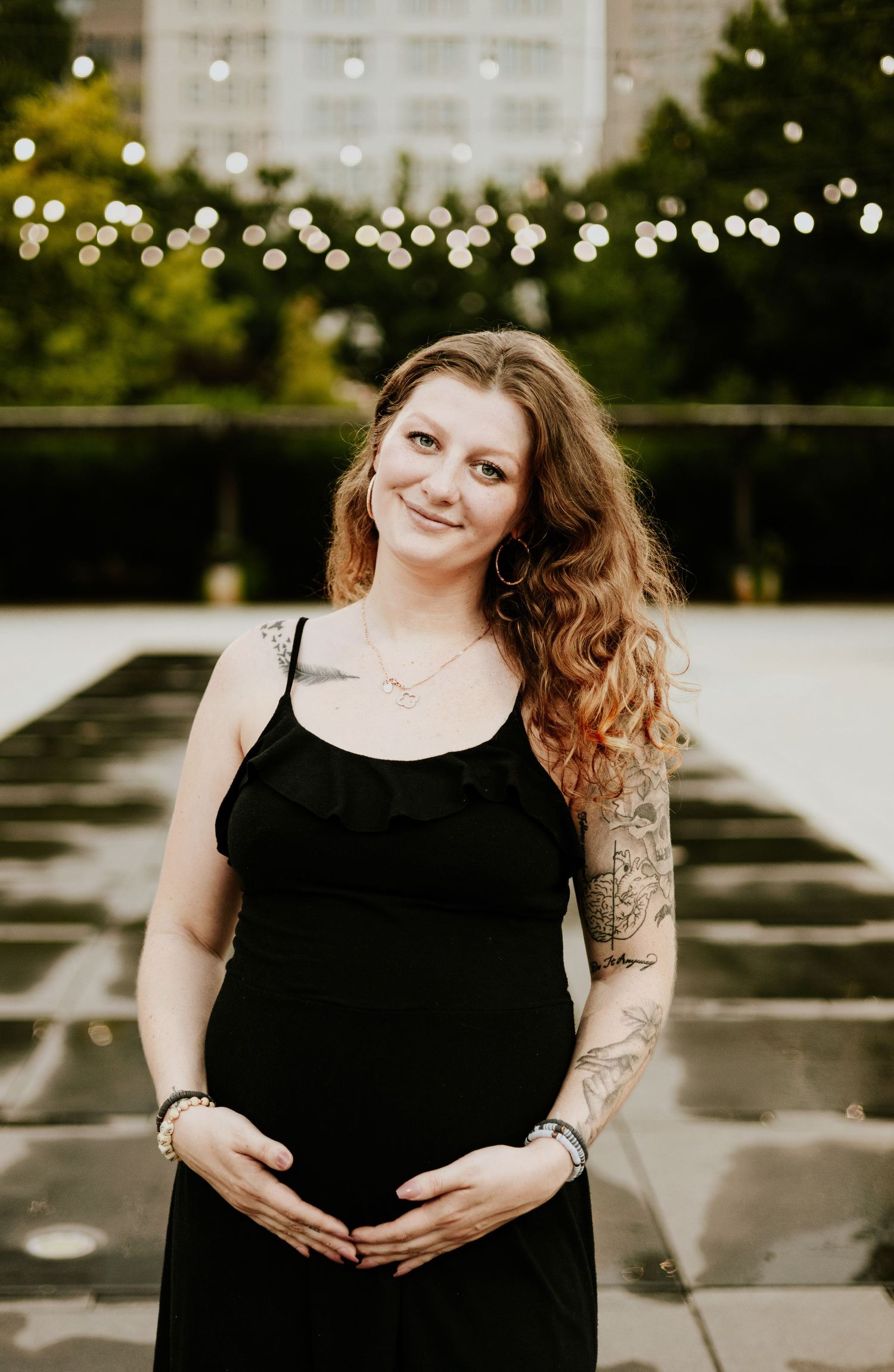 A smiling pregnant woman with tattoos in a black dress cradles her baby bump with string lights behind her.