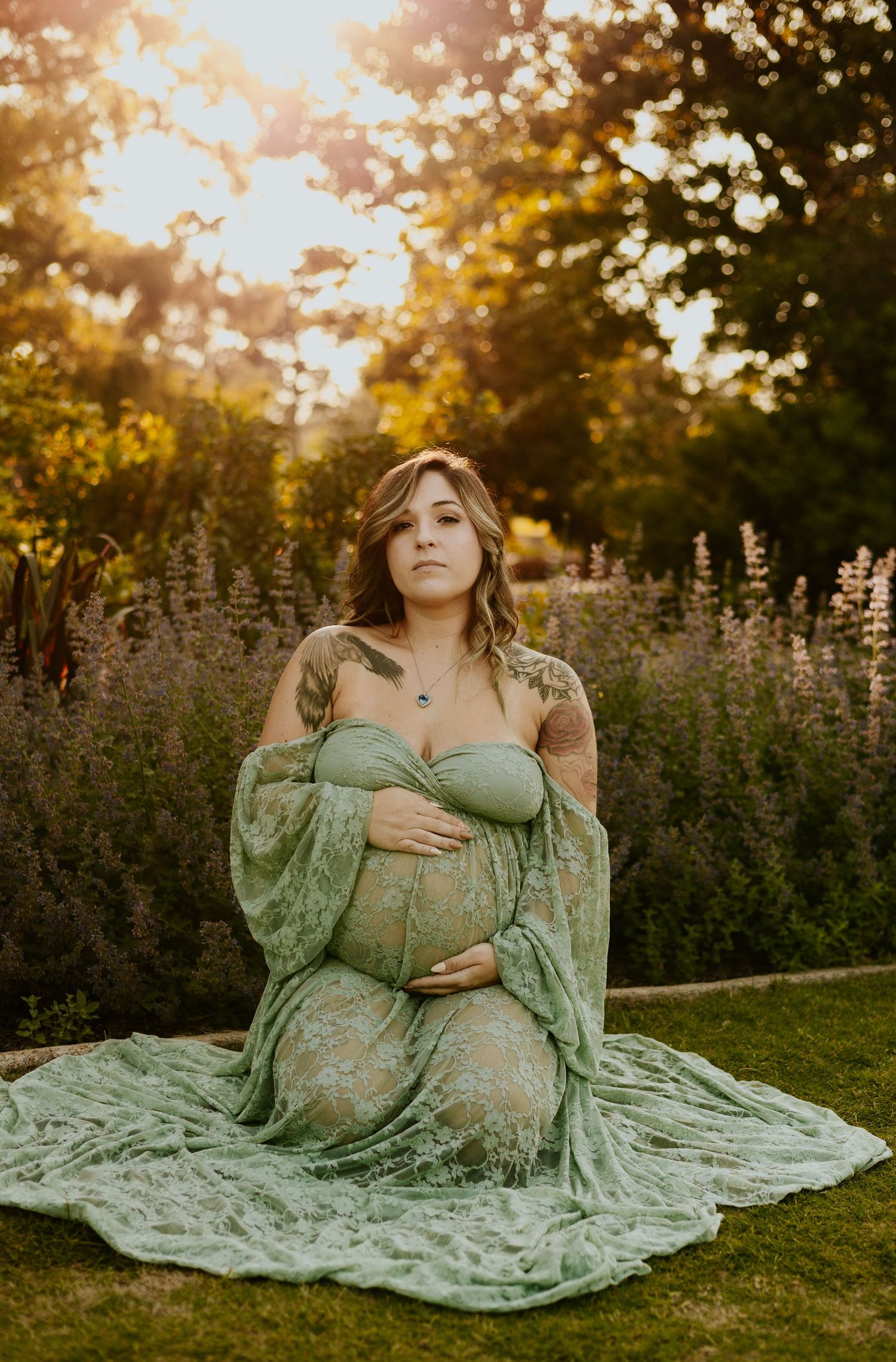 Pregnant woman in sage green lace gown kneeling on grass cradling baby bump surrounded by lavender at sunset.