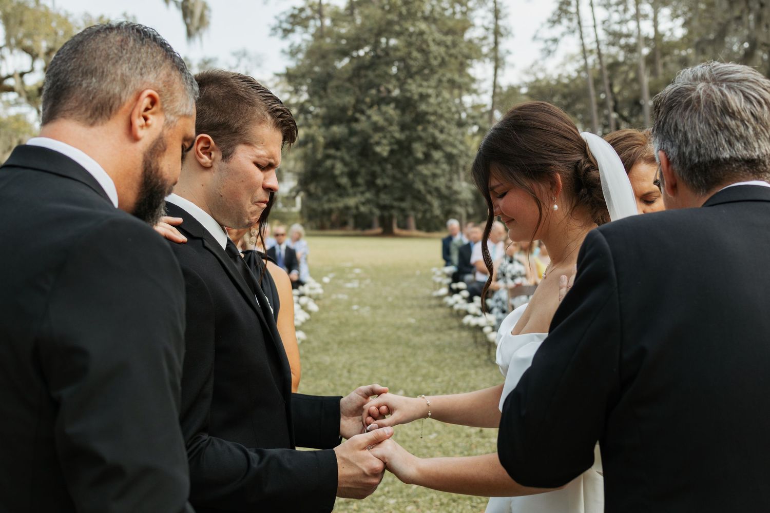 Bride and groom exchanging rings during an outdoor wedding ceremony surrounded by guests in a lush green park setting.