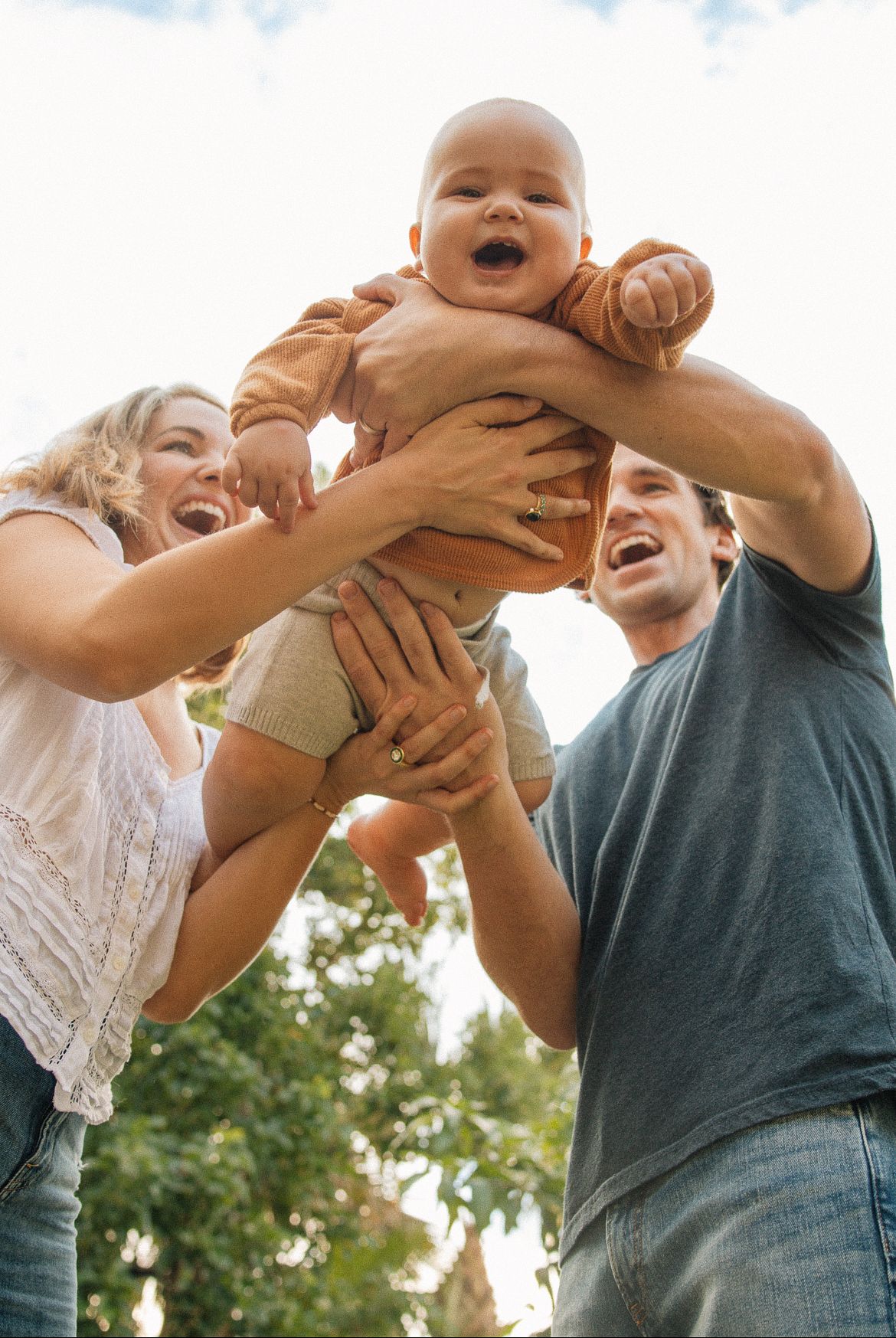Joyful parents lifting laughing baby high in the air outdoors on a sunny day in Palm Beach County, Florida.
