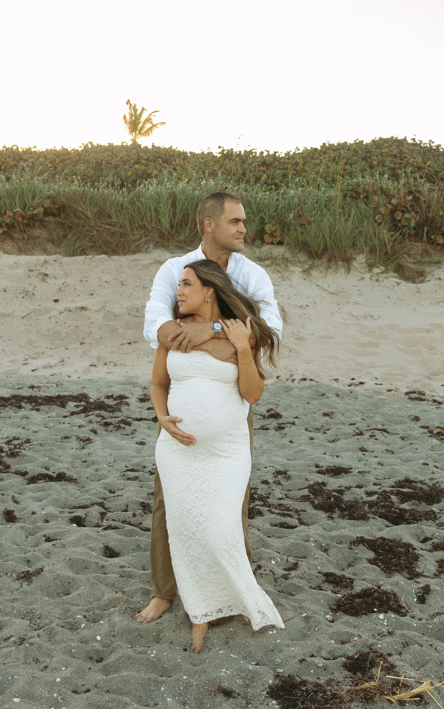 Maternity couple standing together on a beach at dusk, man holding pregnant woman in elegant white lace dress in Palm Beach County, Florida.