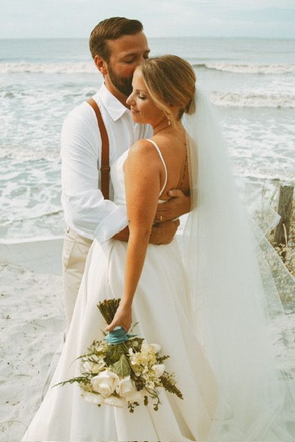 Bride and groom embracing on beach, groom kissing bride holding white bouquet.