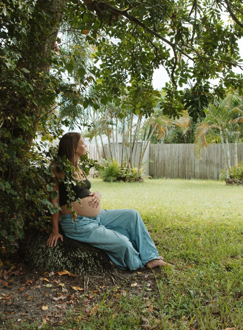 Pregnant woman sitting peacefully under a lush green tree in a tropical backyard, cradling her baby bump.