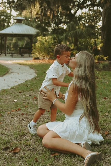 Mother kissing young son in sunny park with gazebo backdrop.