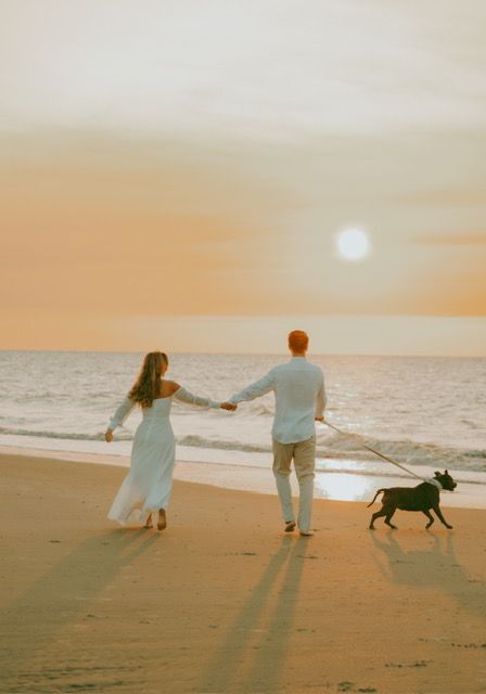 Couple holding hands walking dog on beach at golden sunset.