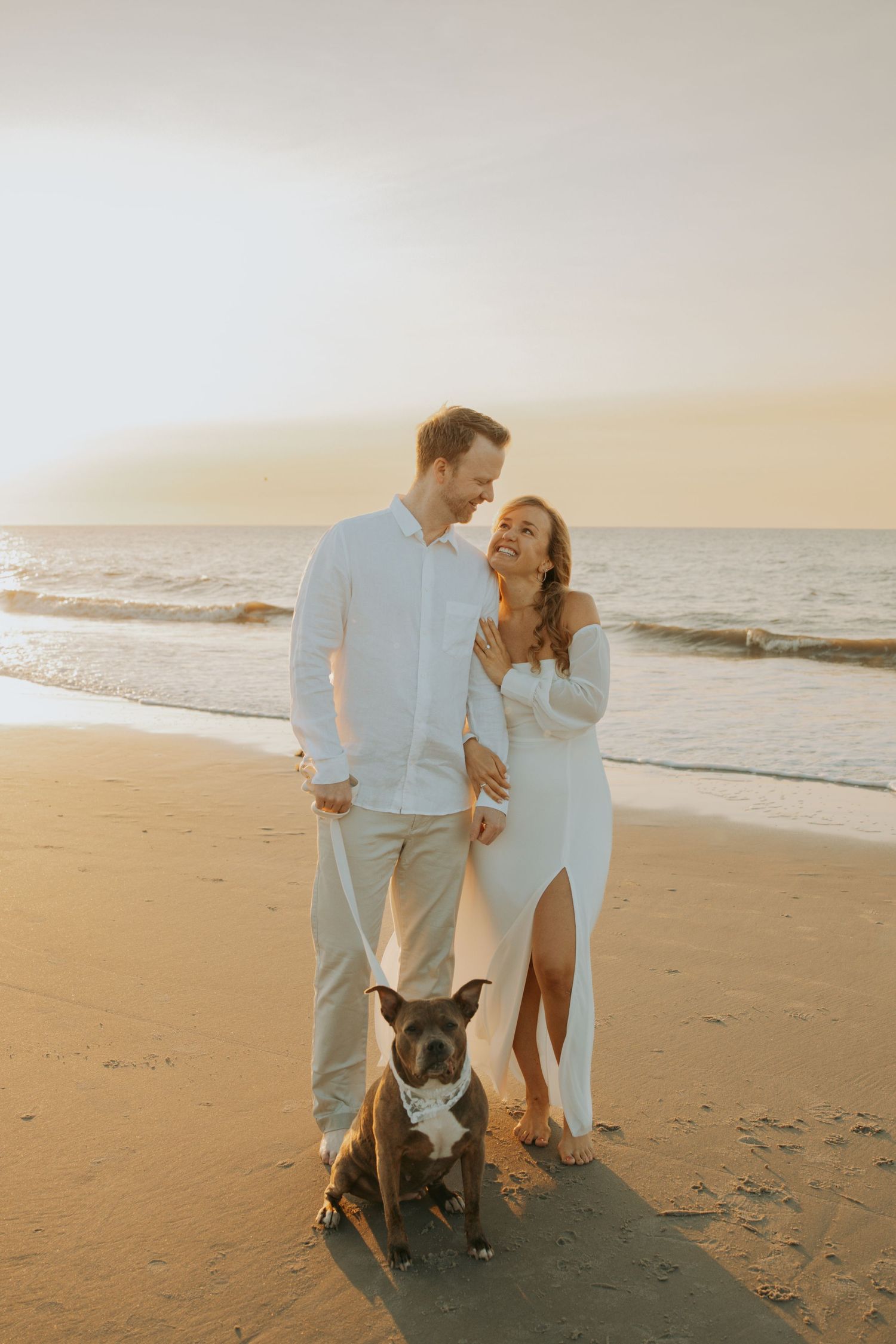 Couple in white outfits with their dog on a sandy beach at golden hour sunrise.