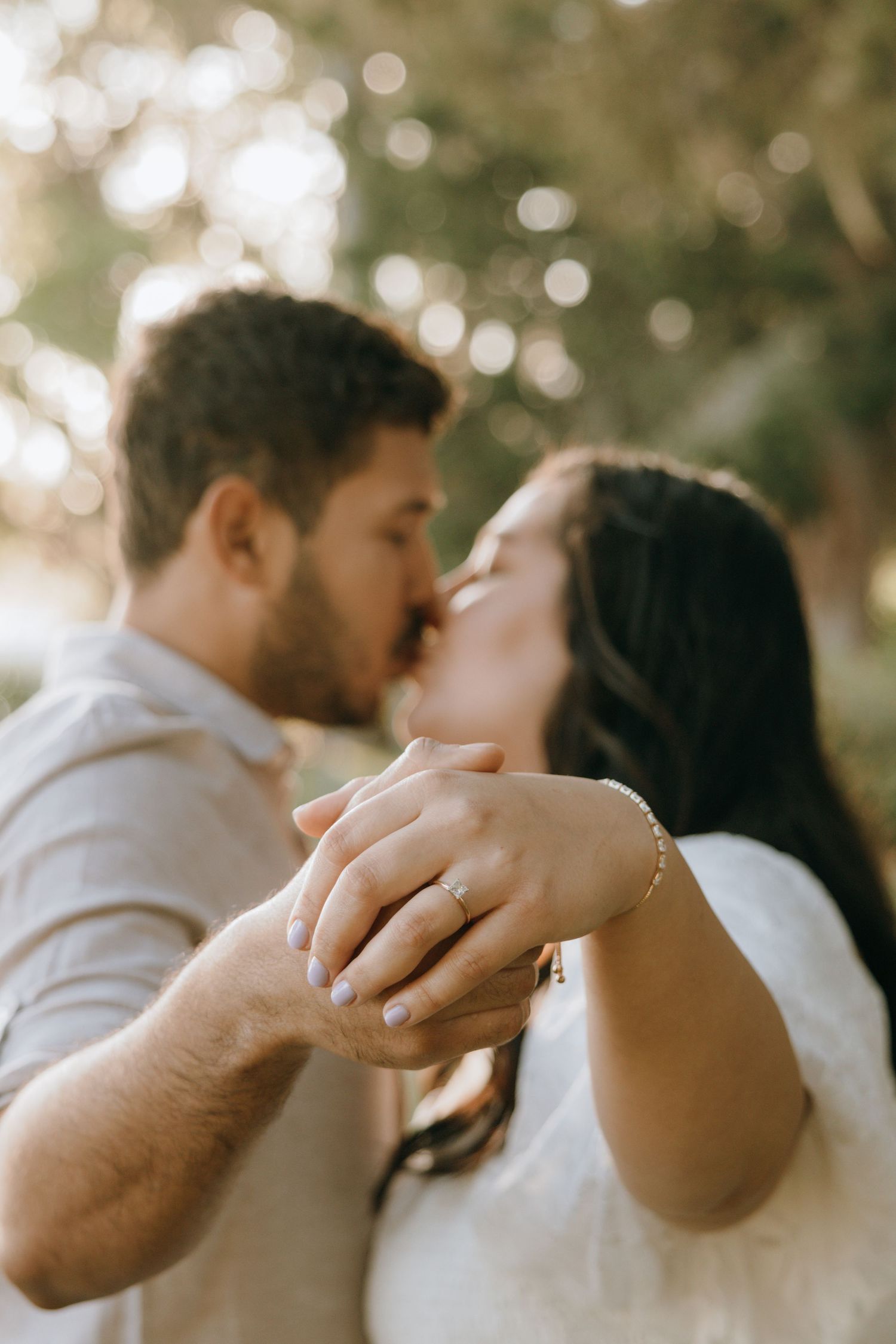 Engaged couple sharing a kiss while holding hands, showcasing a diamond engagement ring in a softly blurred outdoor setting.