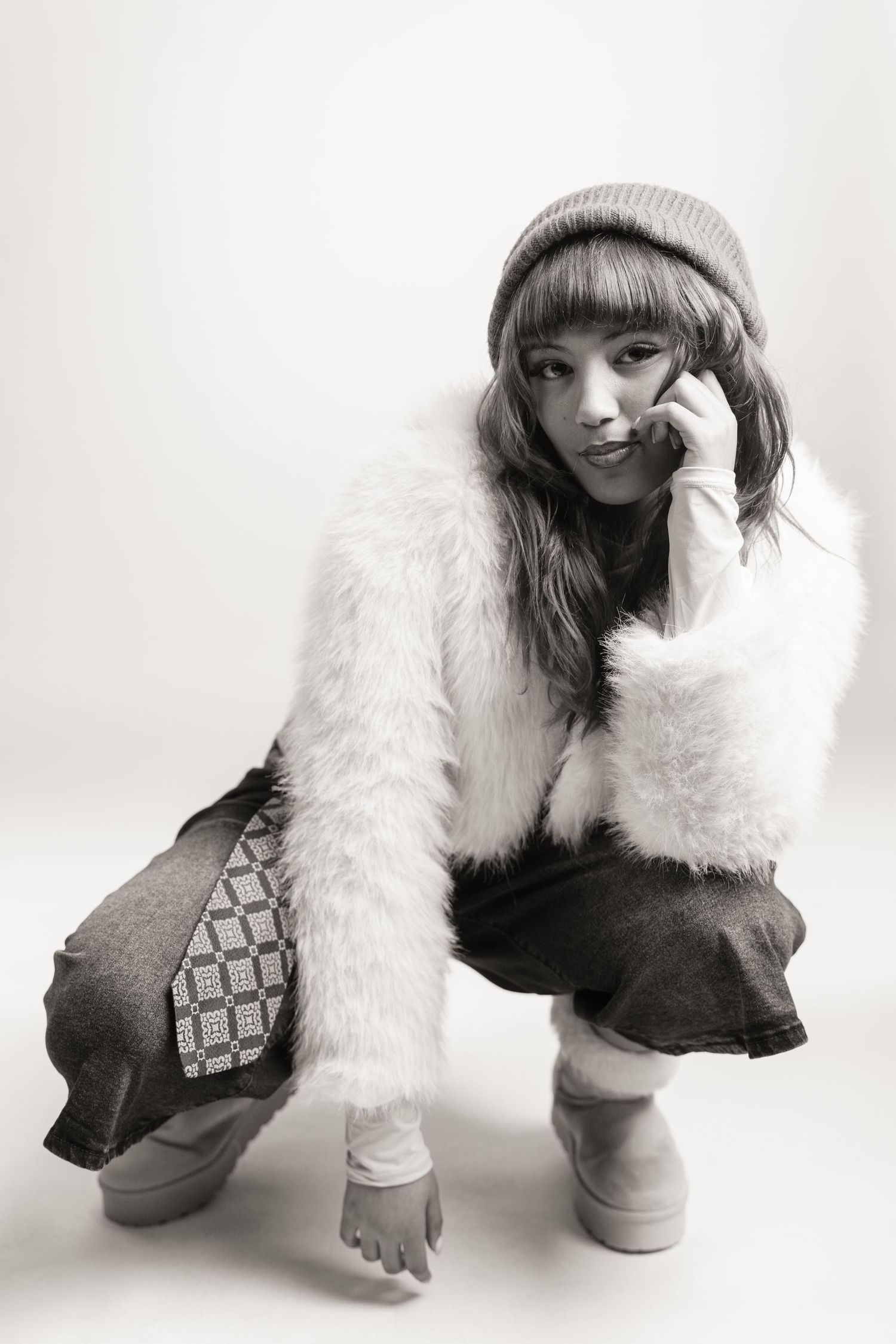 Black and white photo of a young woman crouching in fur vest, beanie hat, and boots in a studio setting.