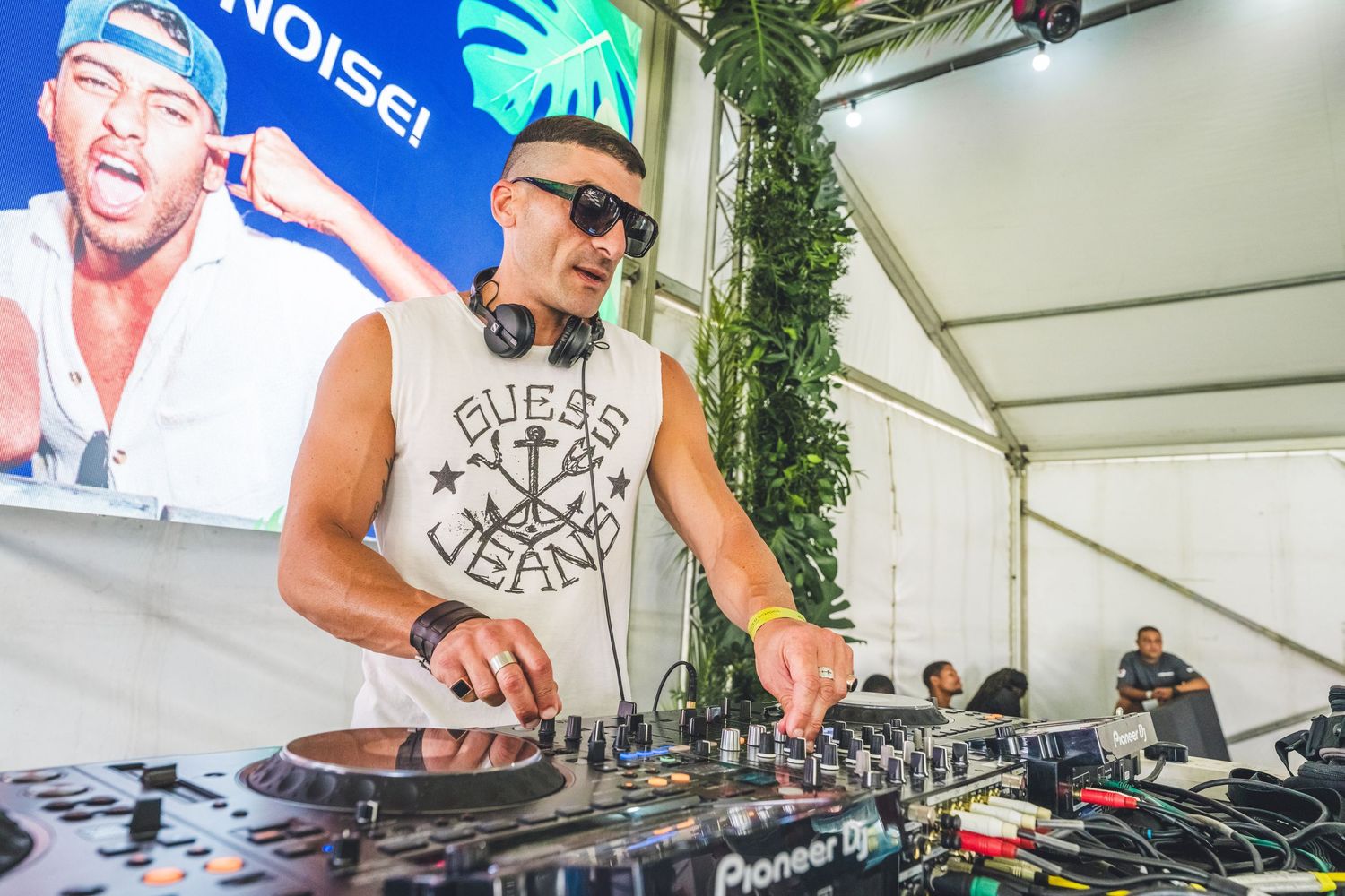 DJ performing at outdoor festival event mixing on Pioneer DJ decks wearing Guess jeans tank top with headphones.