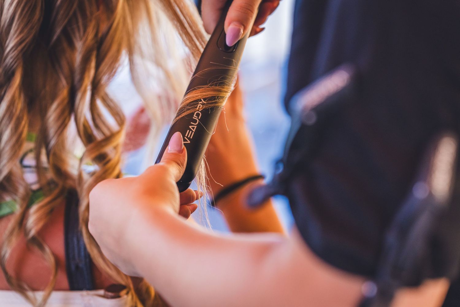 A hairstylist using a curling iron to style long wavy blonde hair in a professional salon setting.