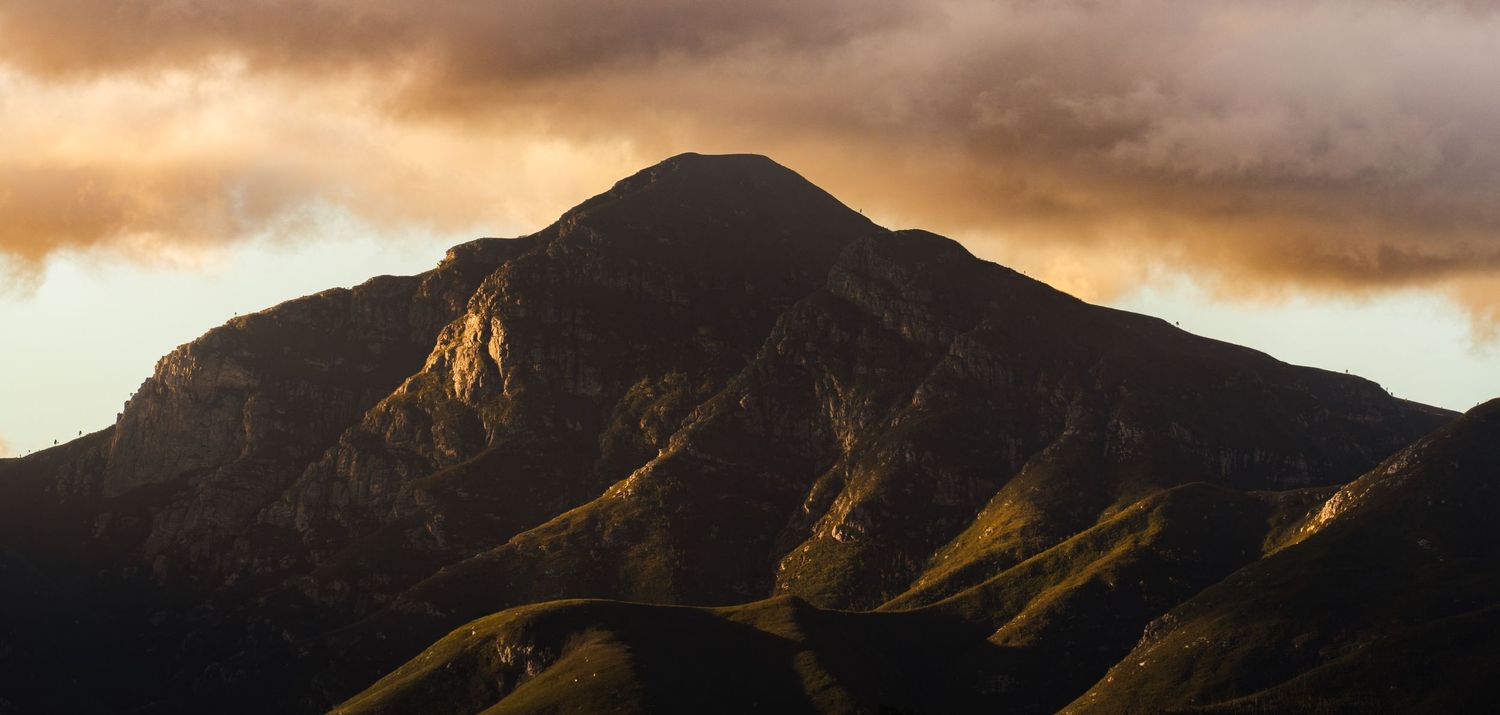 Dramatic mountain peak bathed in golden sunset light with stormy clouds overhead and lush green hills below.
