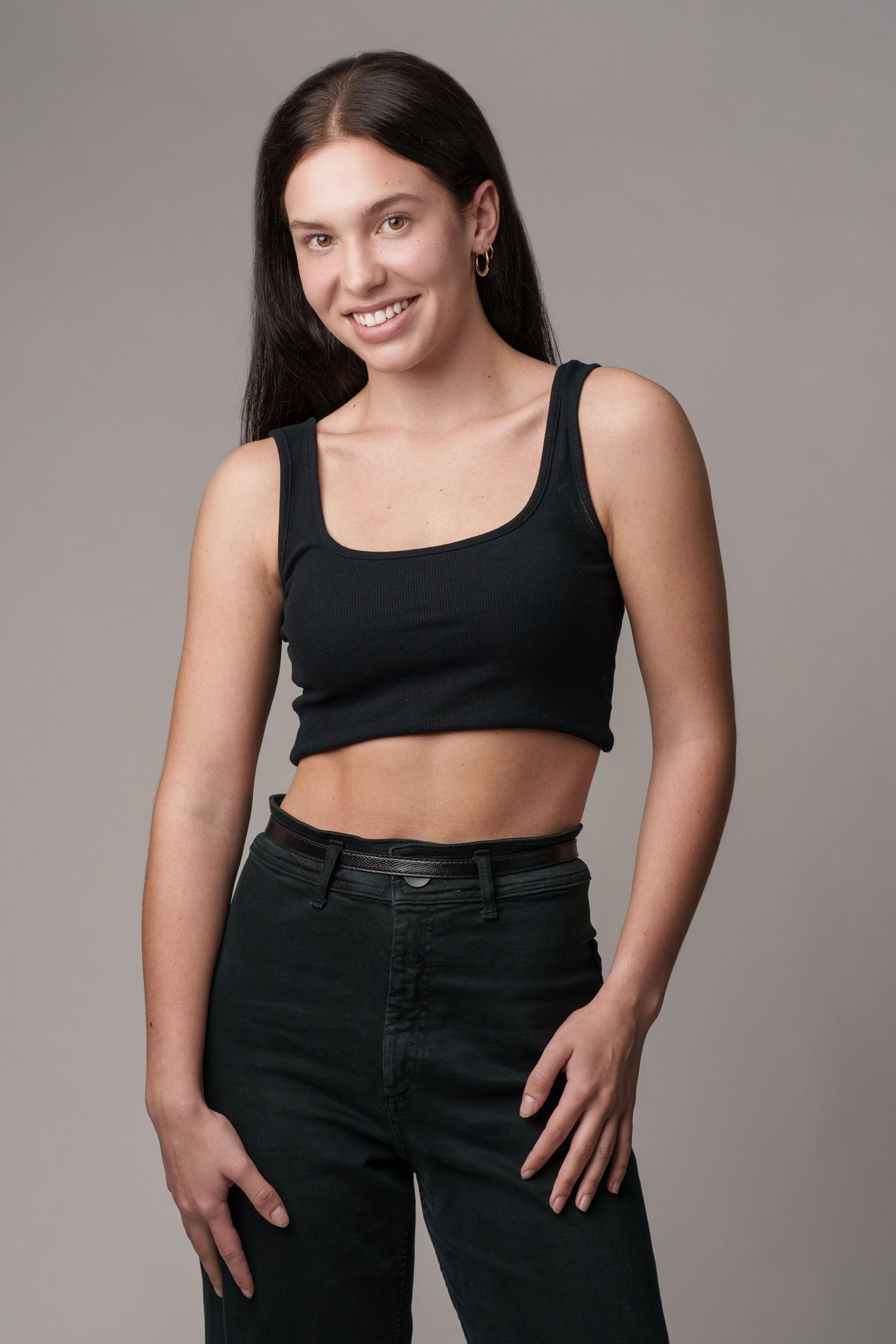 Smiling young woman in black crop top and dark jeans posing against a gray background.