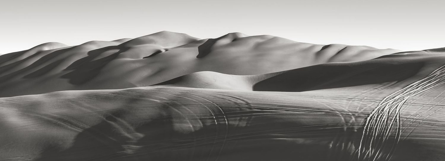 Black and white panoramic photo of sweeping sand dunes with dramatic shadows and wind-swept patterns in a desert landscape.