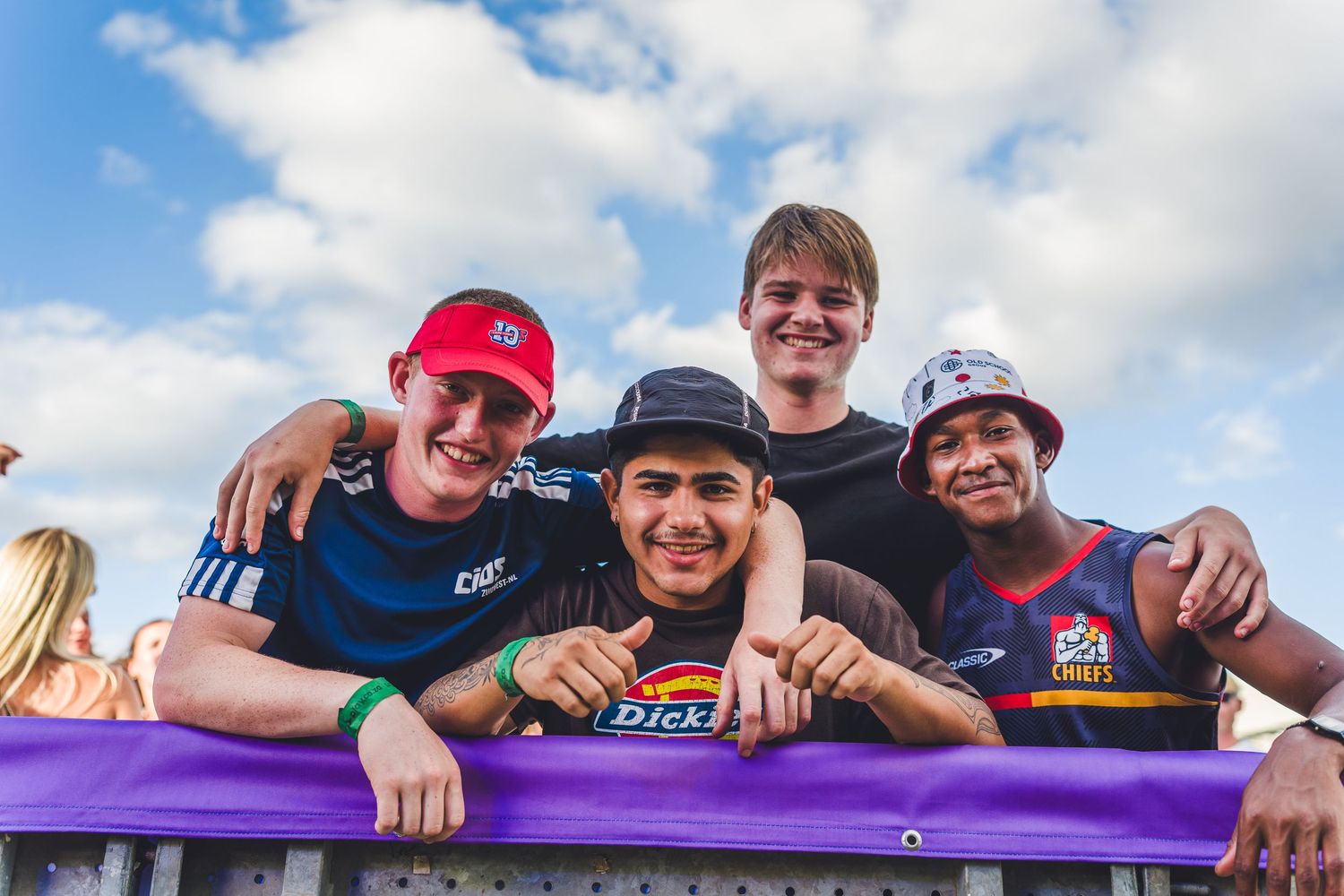 Four smiling young men leaning over a purple barrier at an outdoor festival on a sunny day.