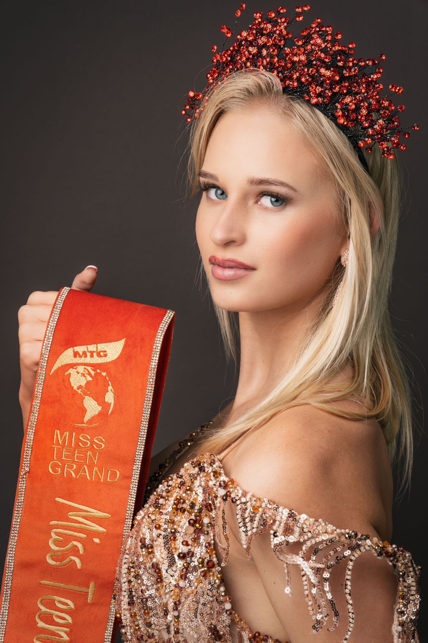Young blonde woman wearing a red jeweled crown and holding a Miss Teen Grand pageant sash in a sparkly gown.