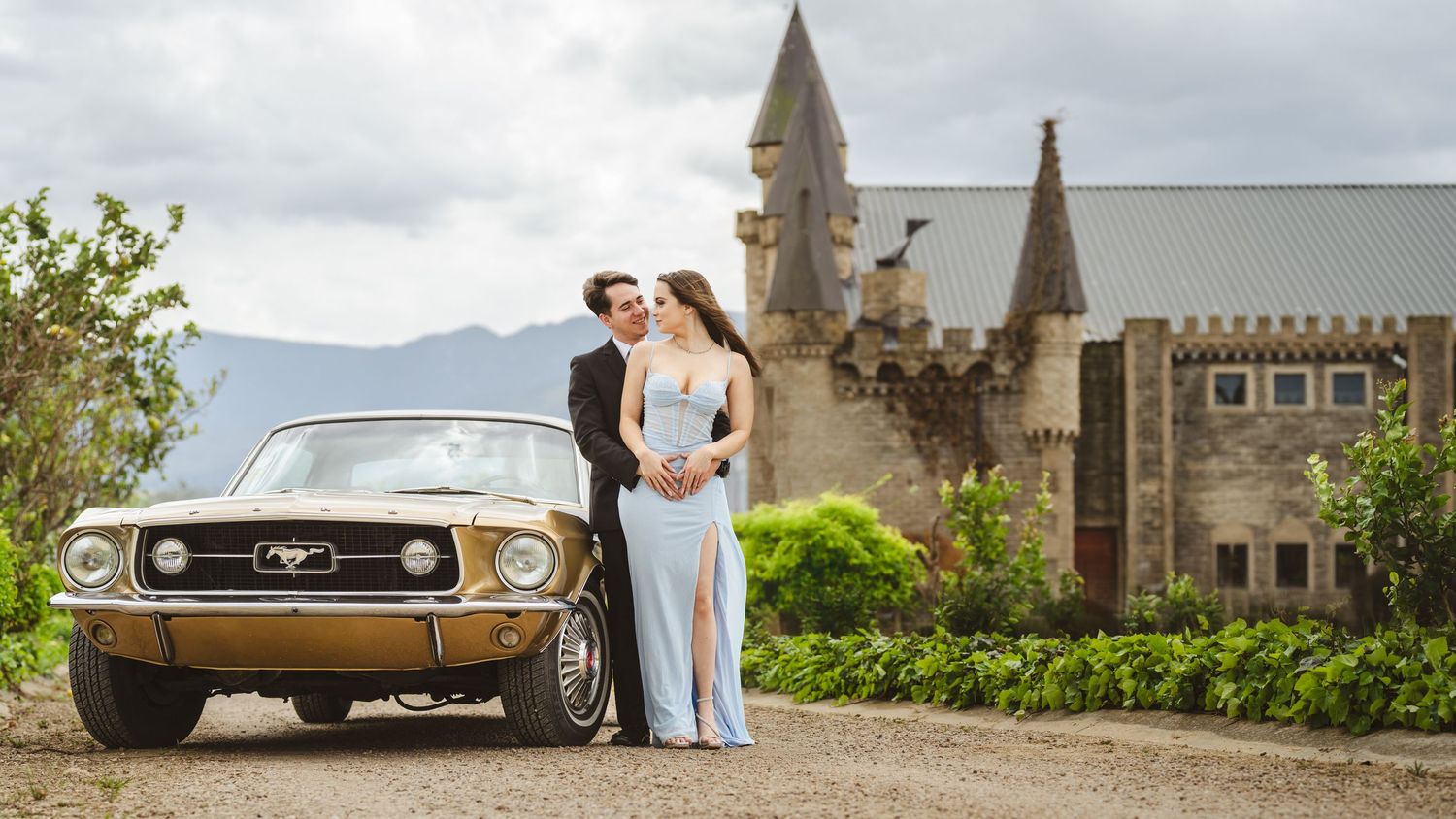 Couple poses by vintage gold Ford Mustang before a castle backdrop with mountain views.