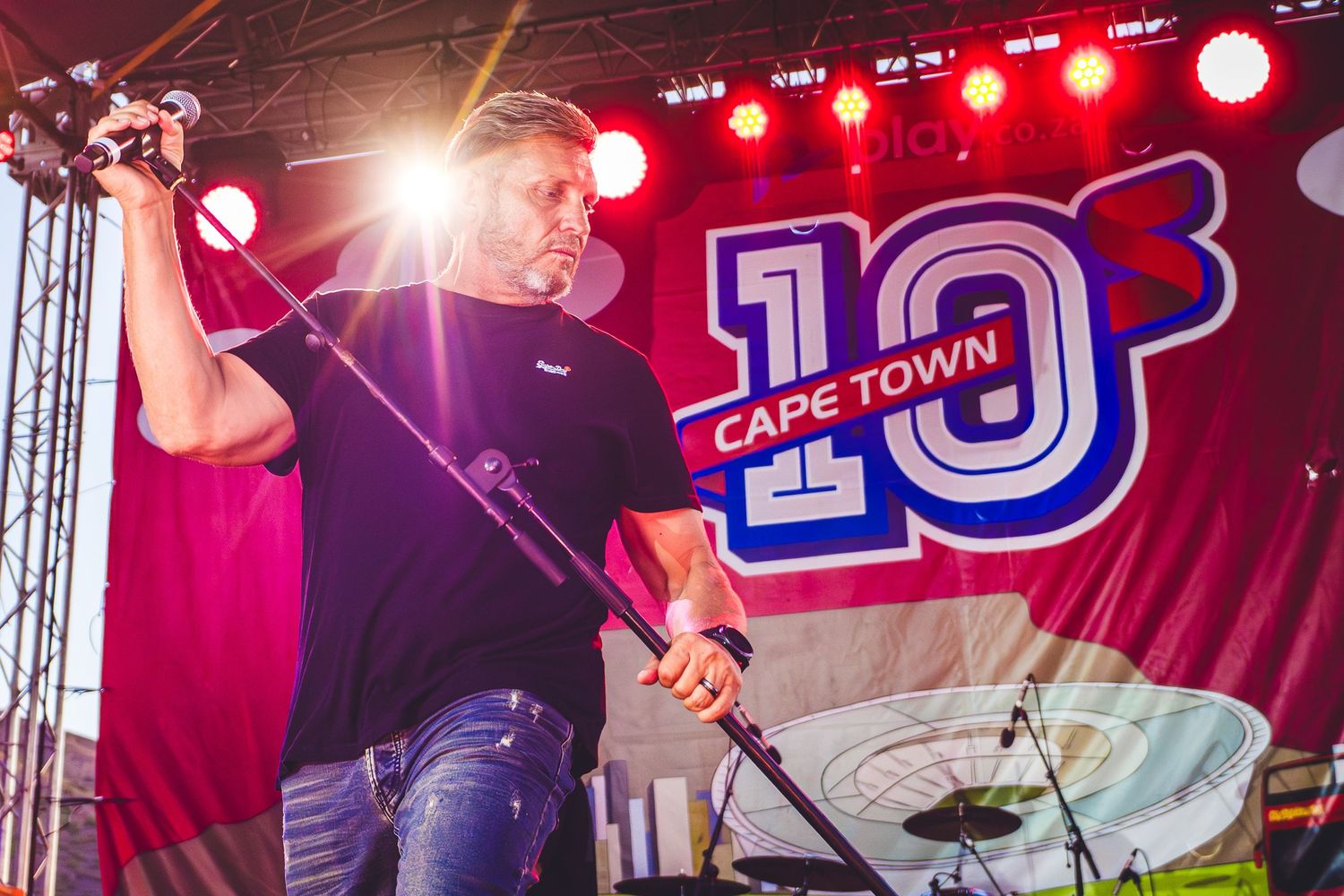 Man performing on stage at Cape Town 10s event, holding microphone stand under colorful lights with branded banner behind.