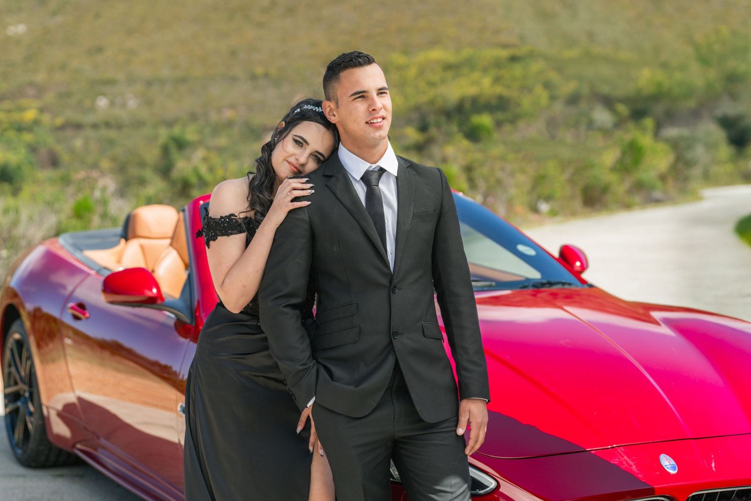 A couple poses with a red Ferrari sports car on a scenic mountain road.