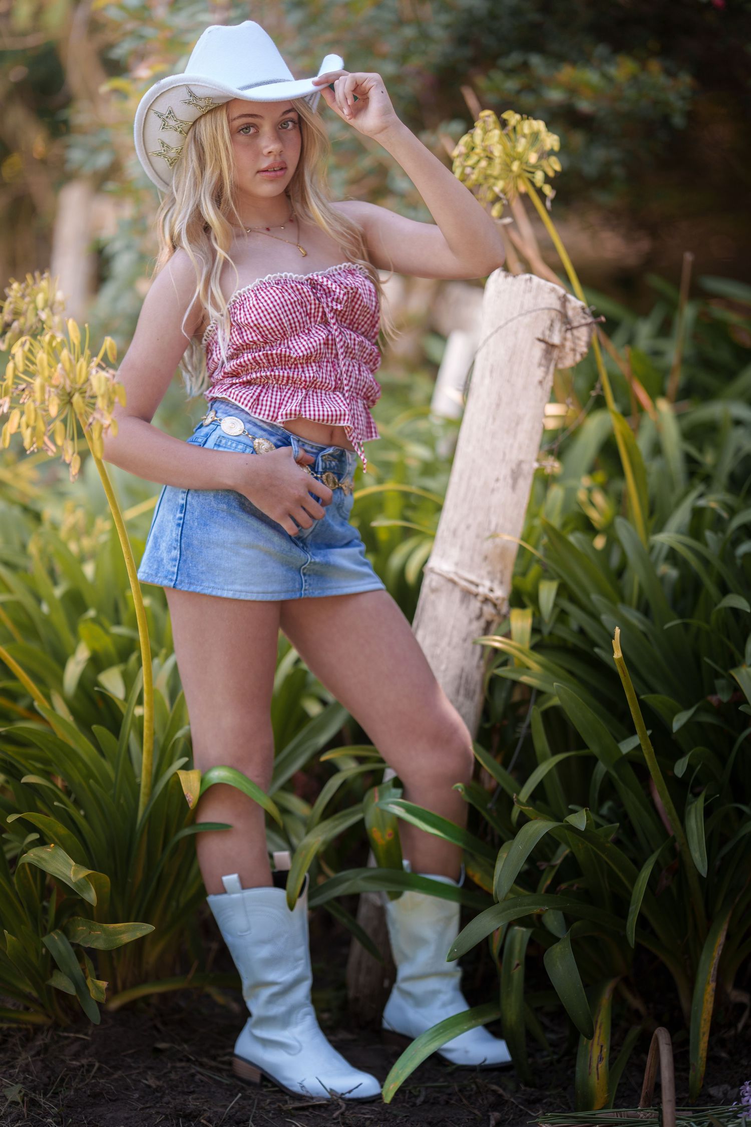 A person in denim shorts and white cowboy boots poses among lush green plants in a garden setting.