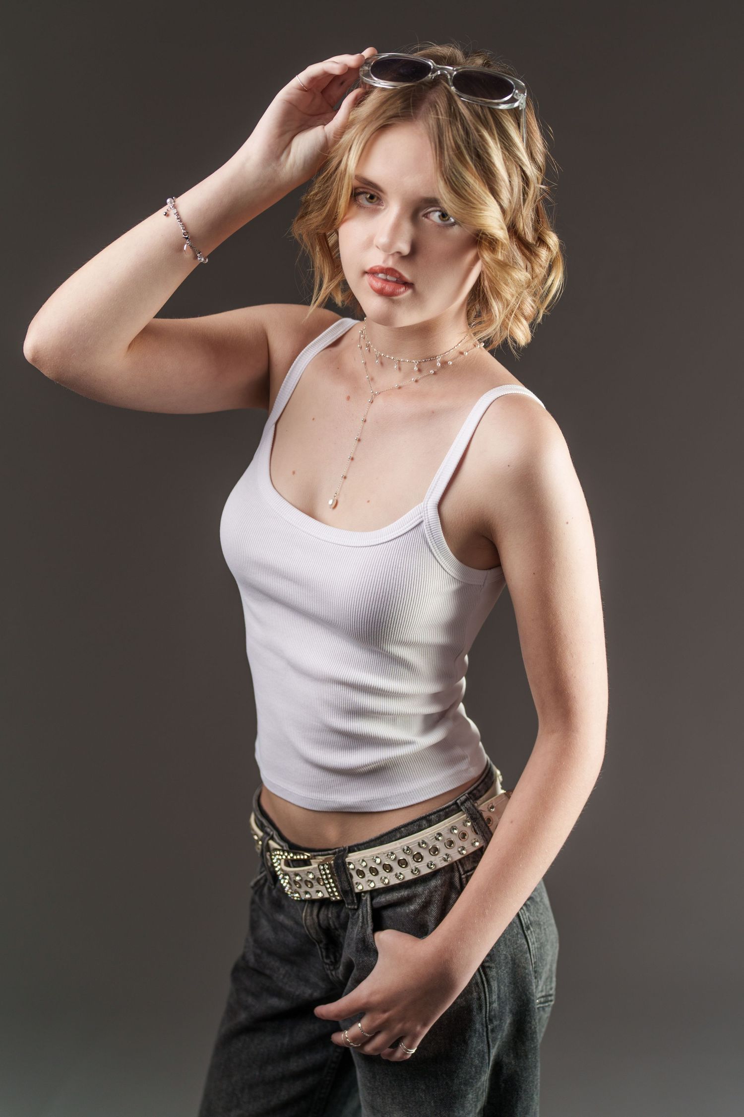 Young woman in white tank top and jeans posing with sunglasses on head against gray background.